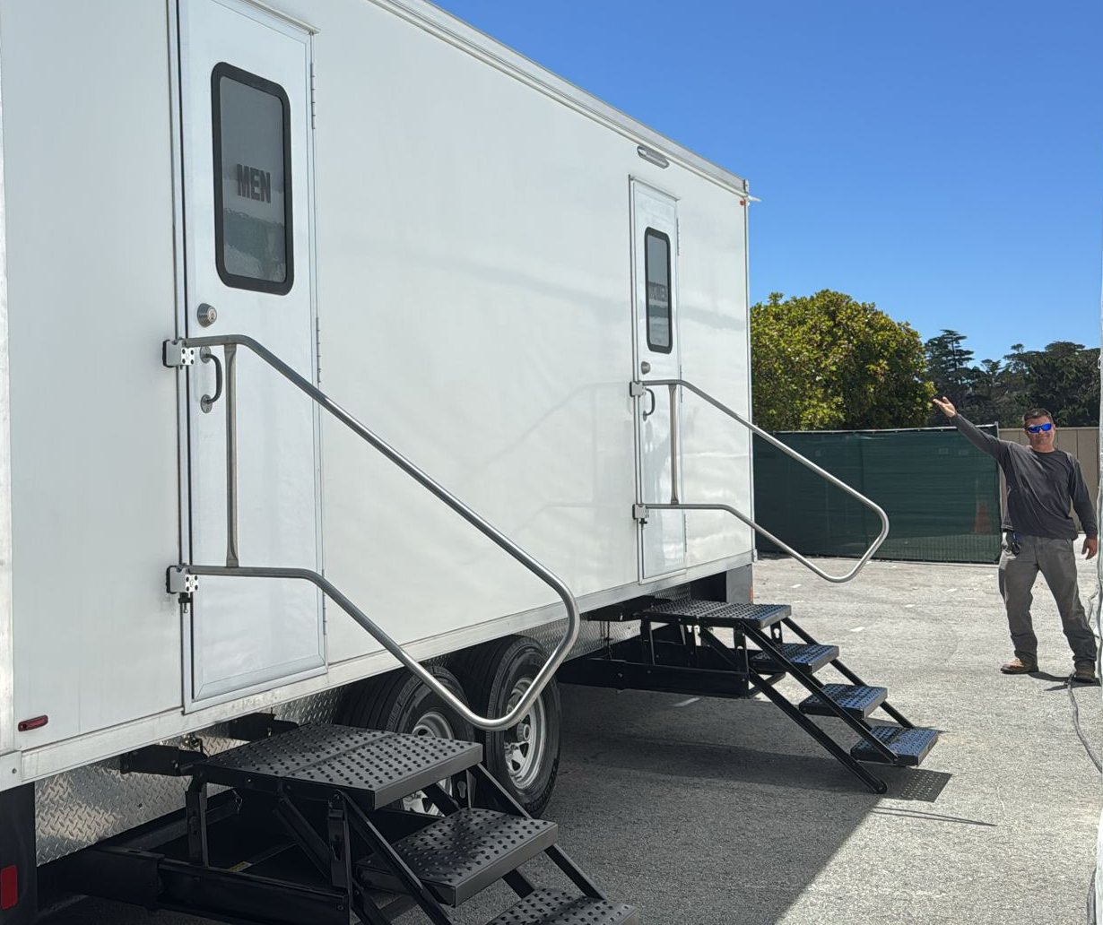 A crew member points to a Lavatory restroom trailer set up with steps and stainless handrails at each door