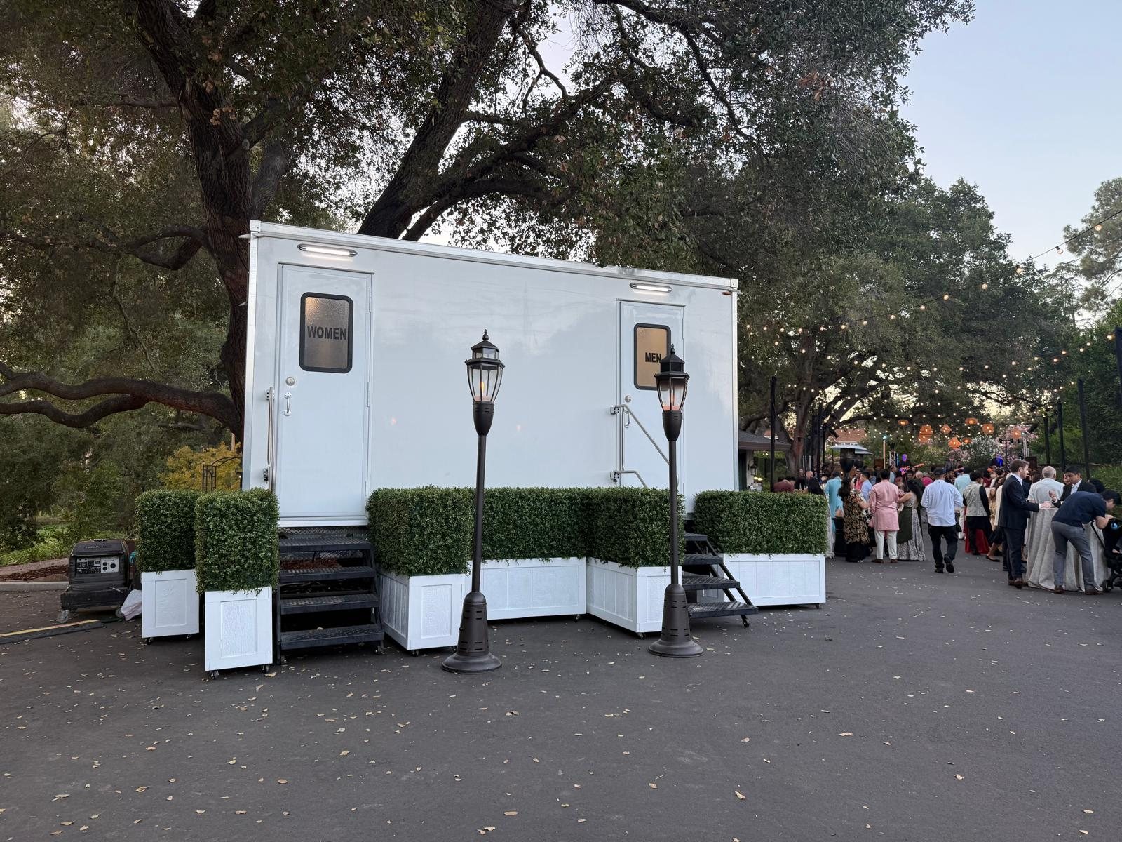 Wedding guests gather under string lights beside a Lavatory restroom trailer dressed with hedge planters and lamp posts
