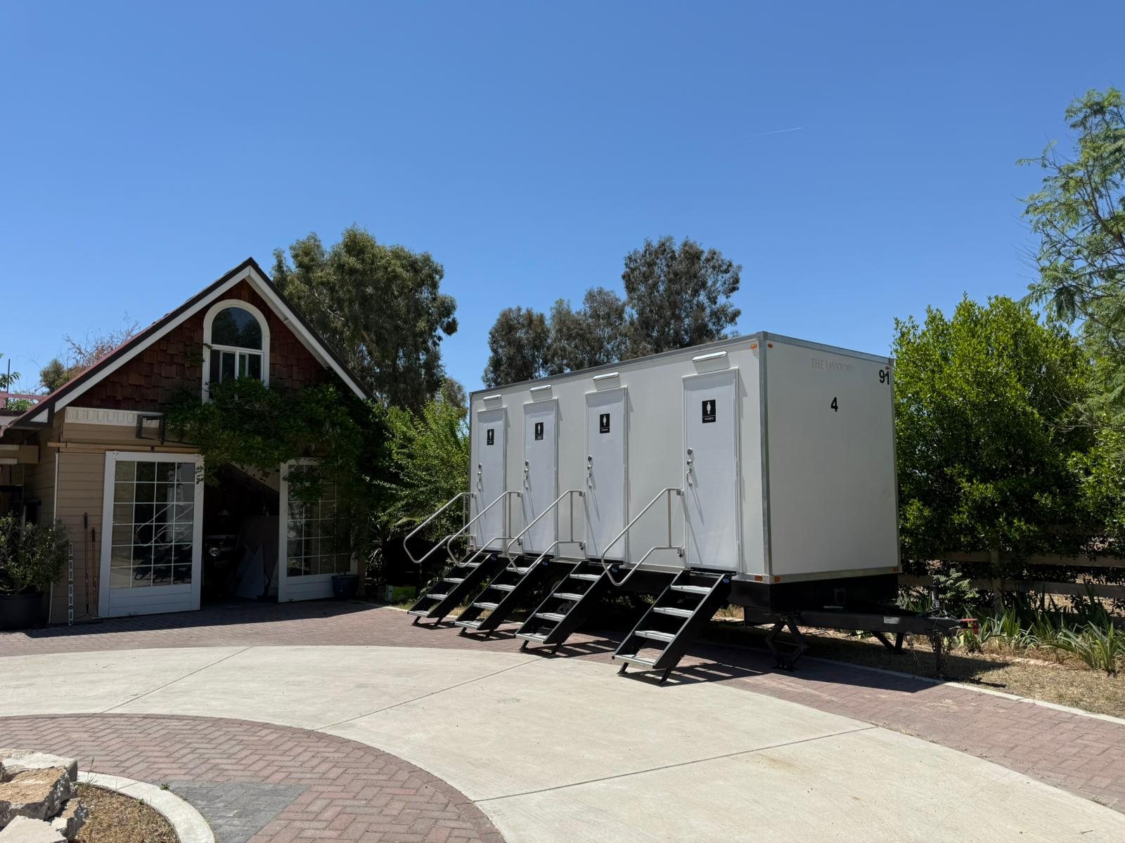 A Lavatory 4-Station luxury restroom trailer parked on a paved circle drive beside a cottage-style venue building in Clovis