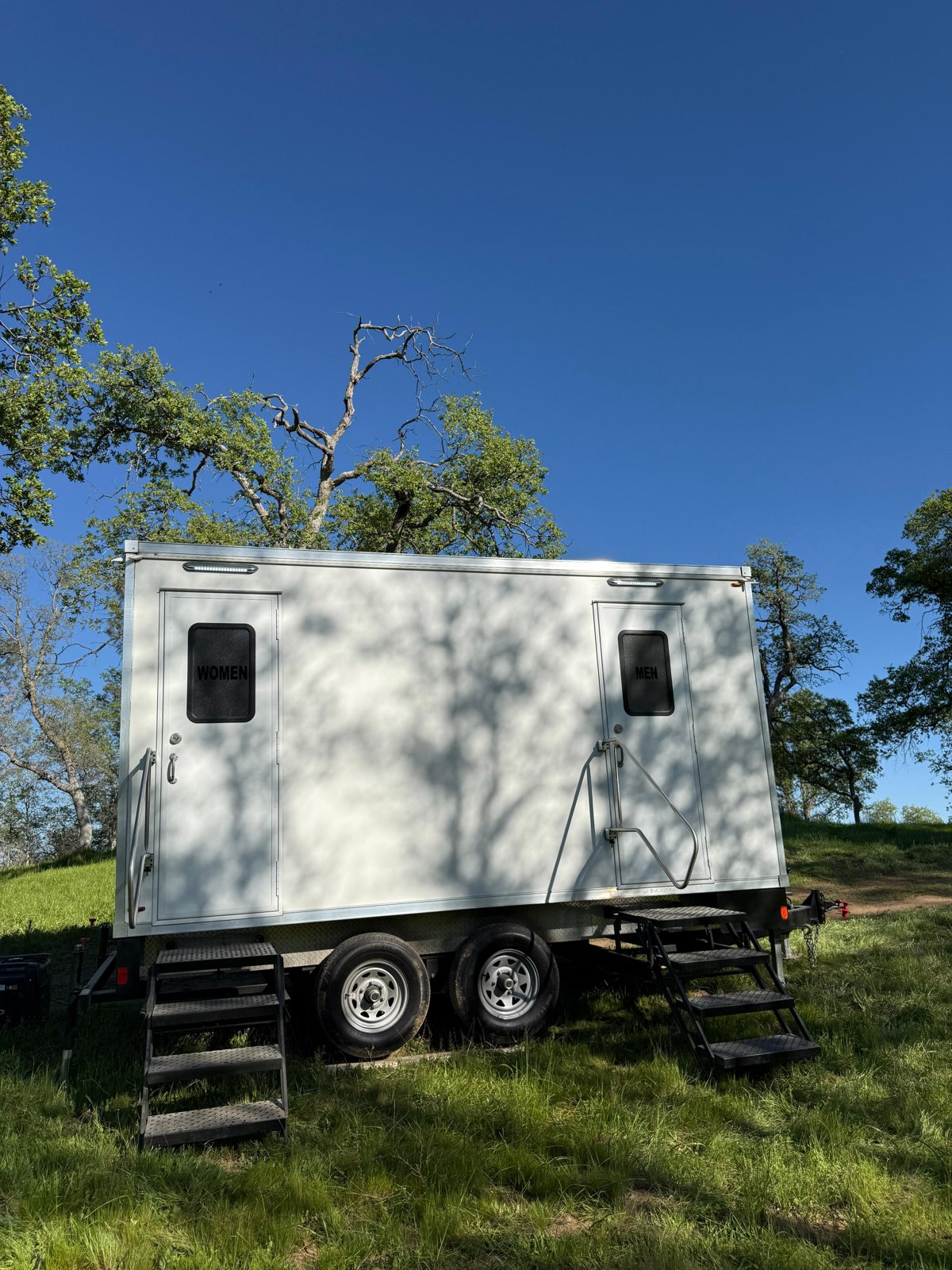 A Lavatory two-room restroom trailer with women's and men's doors on a grassy hillside under oak trees
