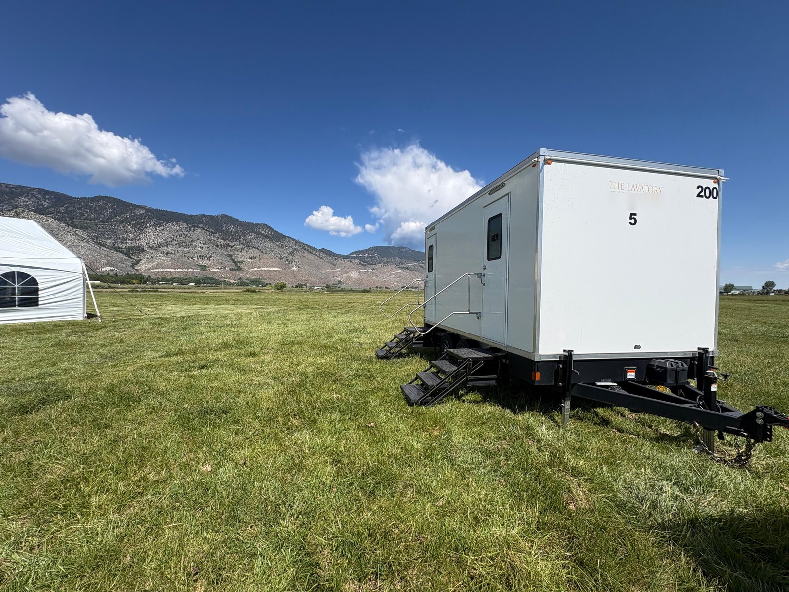 A Lavatory 5-Station luxury restroom trailer on a mowed grass field beside a white event tent, with dry mountains behind