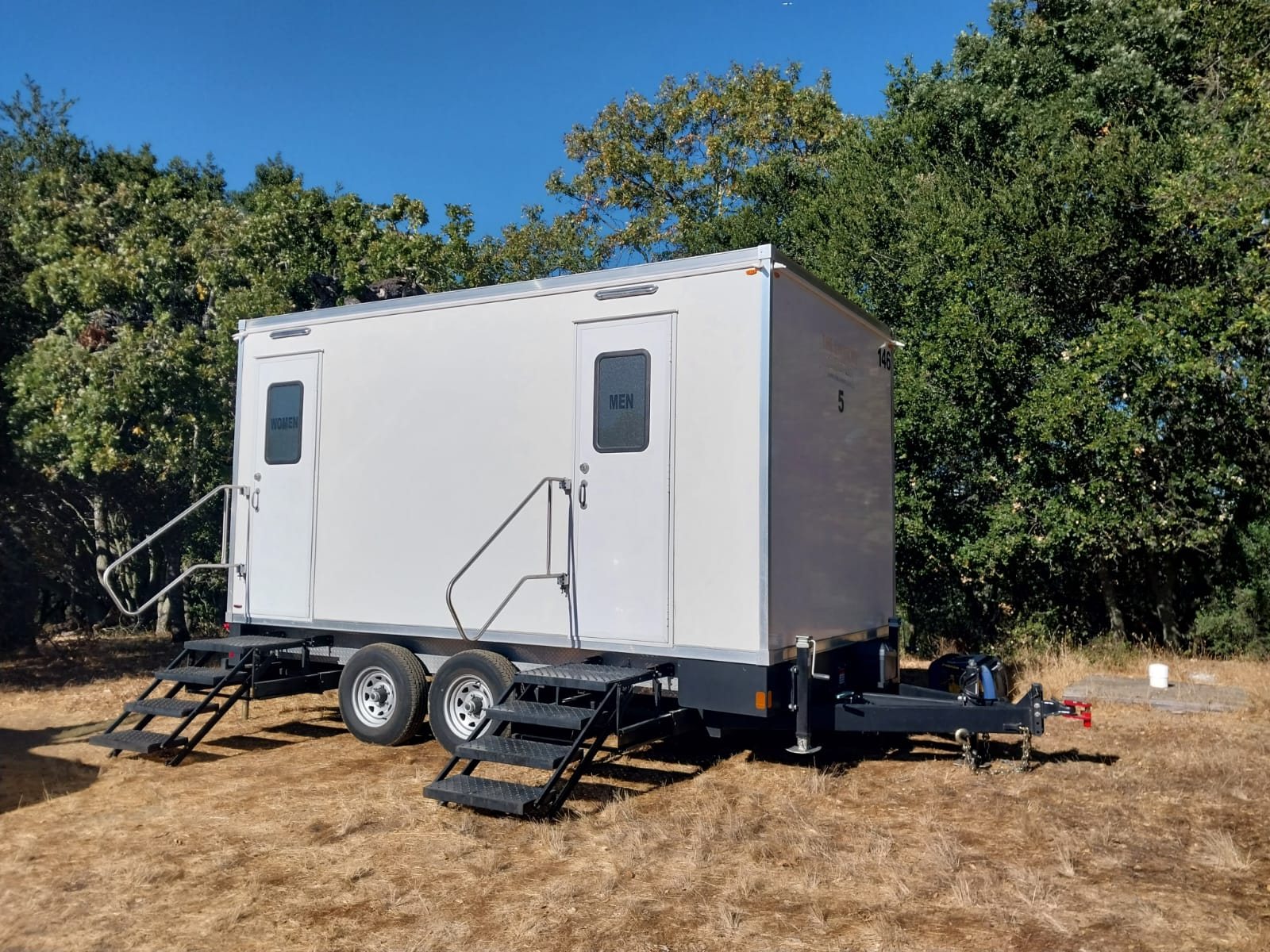 A Lavatory 5-Station restroom trailer on dry grass in front of a line of green oak trees