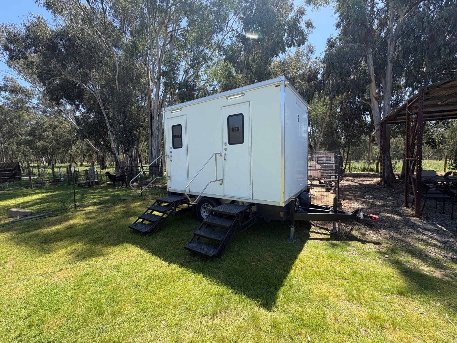 A Lavatory 2-Station restroom trailer on a green lawn at a ranch property, with tall eucalyptus trees and a wooden shade structure behind it