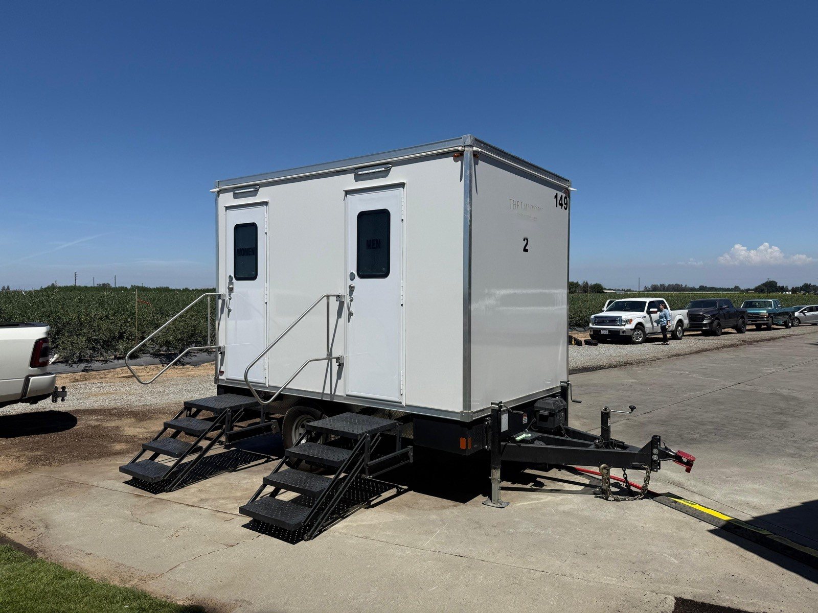 A Lavatory 2-Station restroom trailer on a paved lot beside open farm fields, with pickup trucks parked behind it