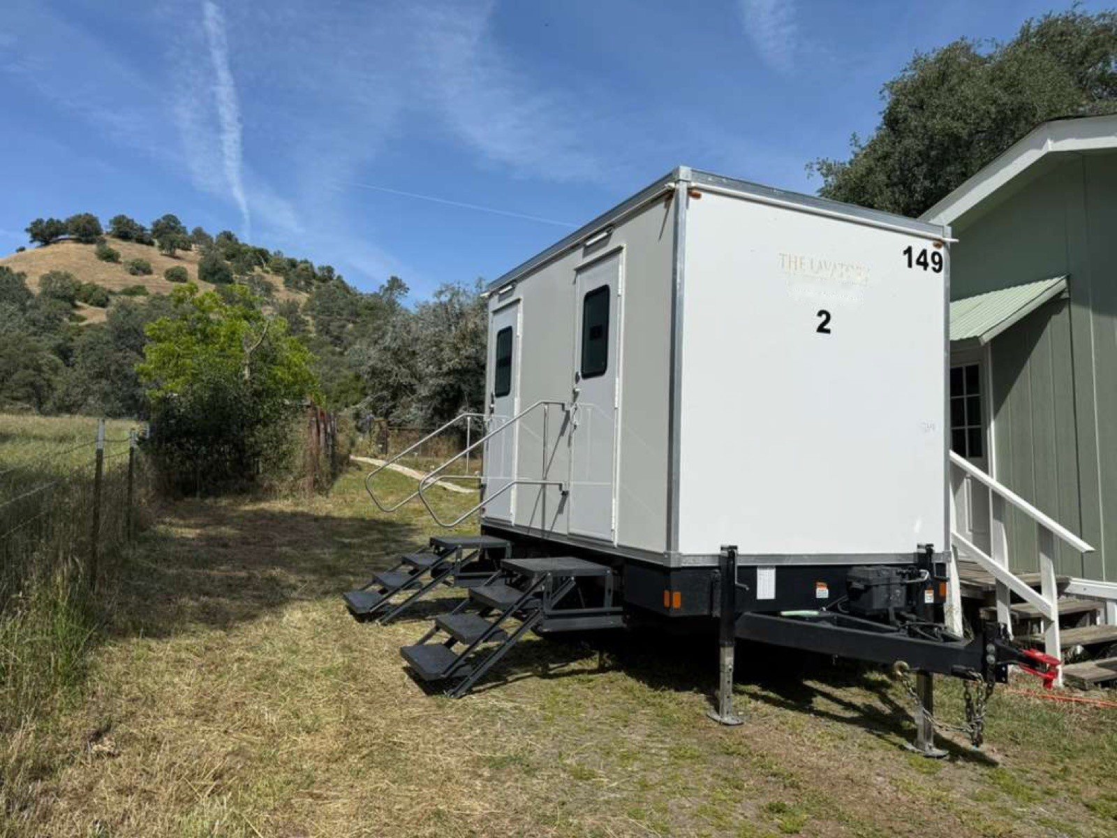 A Lavatory 2-Station restroom trailer on grass beside a small house, with oak-covered hills behind it