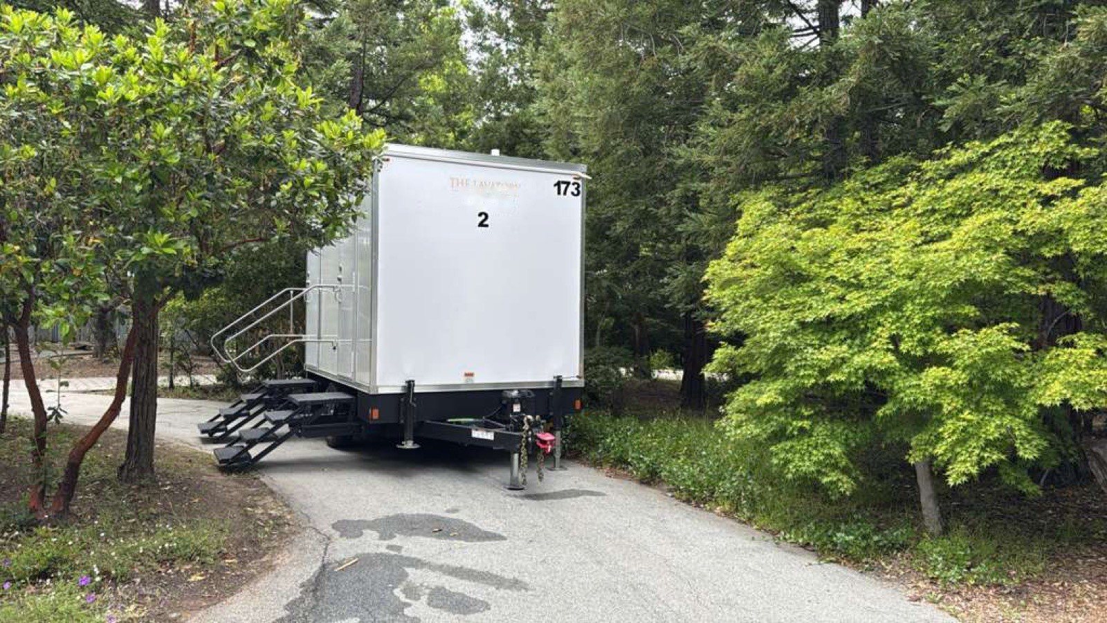 A Lavatory 2-Station restroom trailer parked on a narrow paved drive among leafy trees