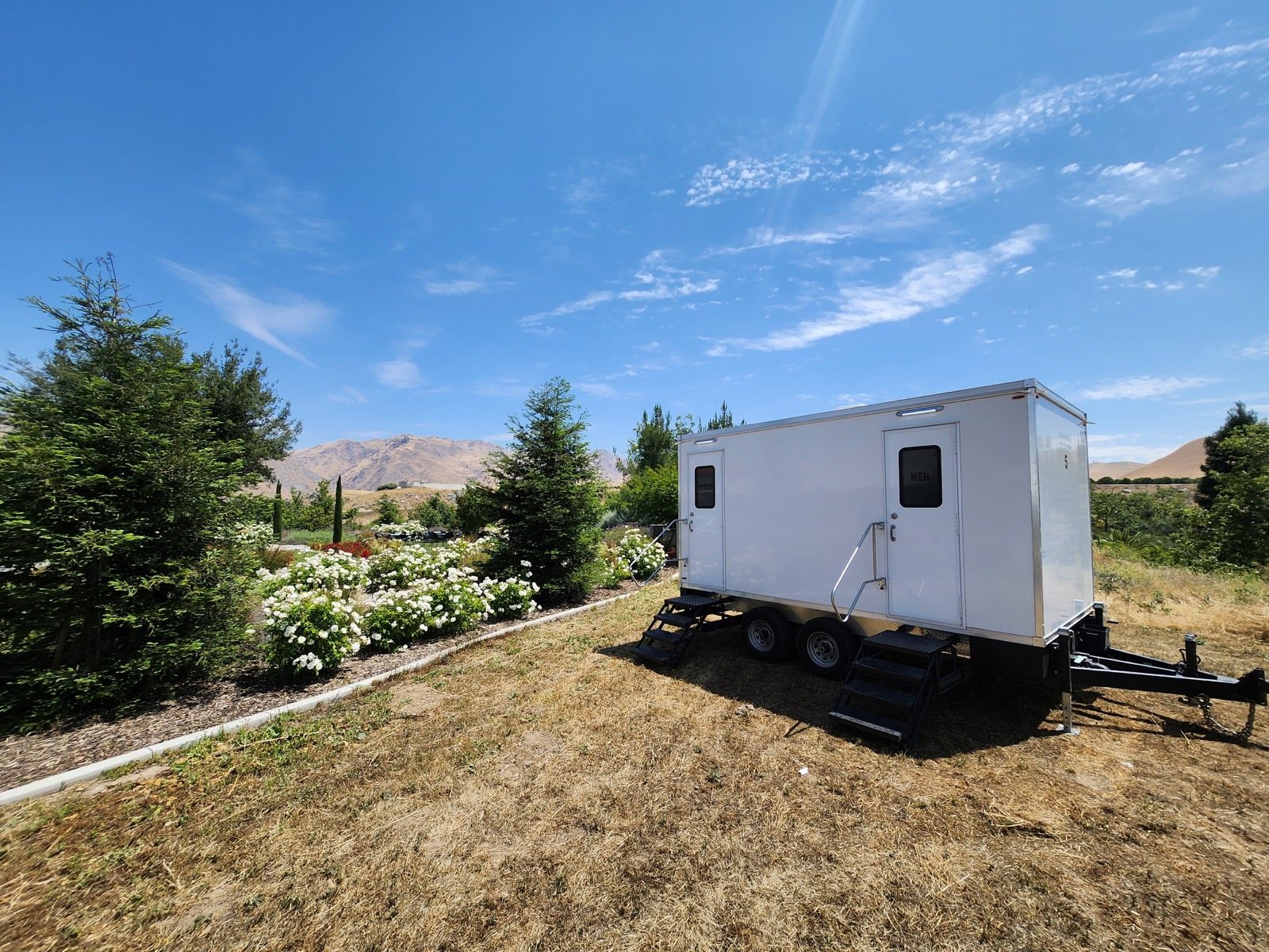 A Lavatory restroom trailer on dry grass beside a garden bed of white hydrangeas, with mountains in the distance