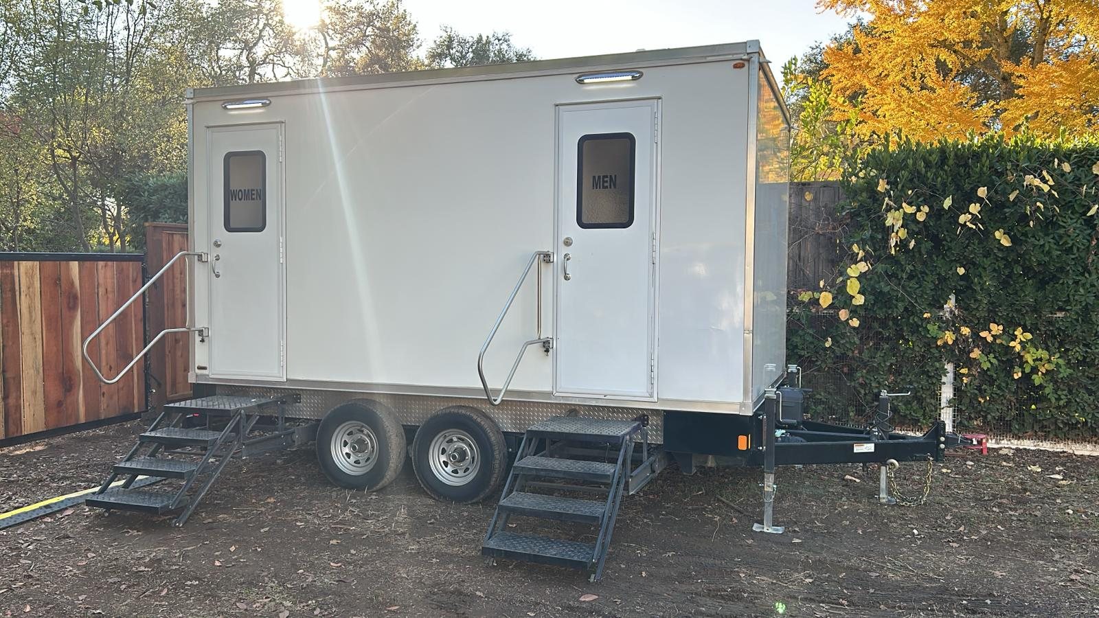 A Lavatory 5-Station restroom trailer set up in a backyard beside a wood fence and a yellow fall tree