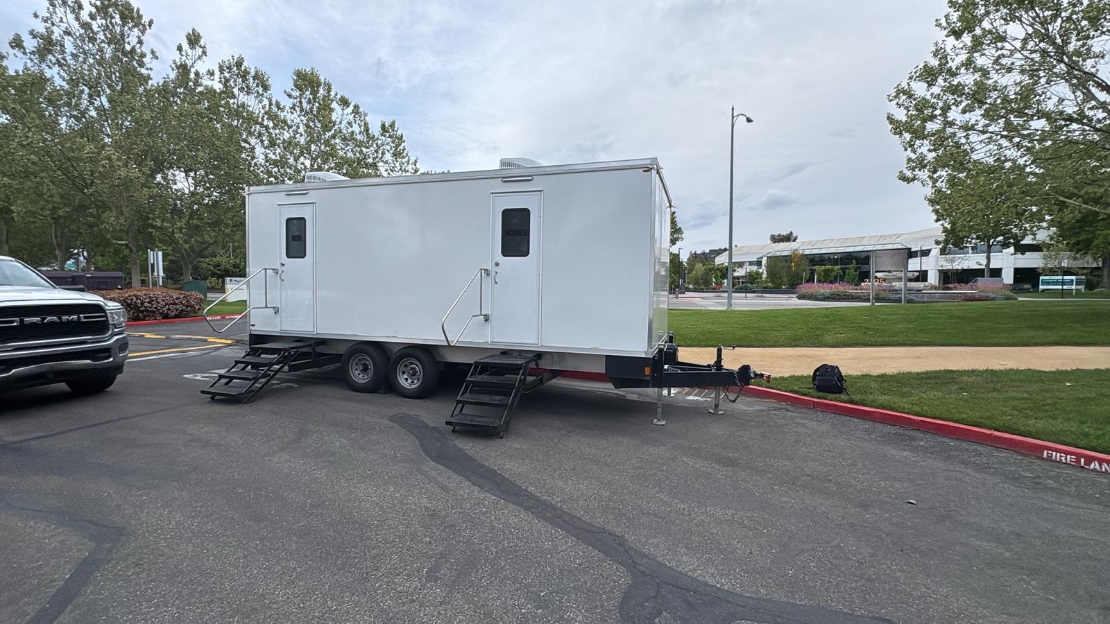 A Lavatory restroom trailer in an office park parking lot, with a low office building and trees in the background