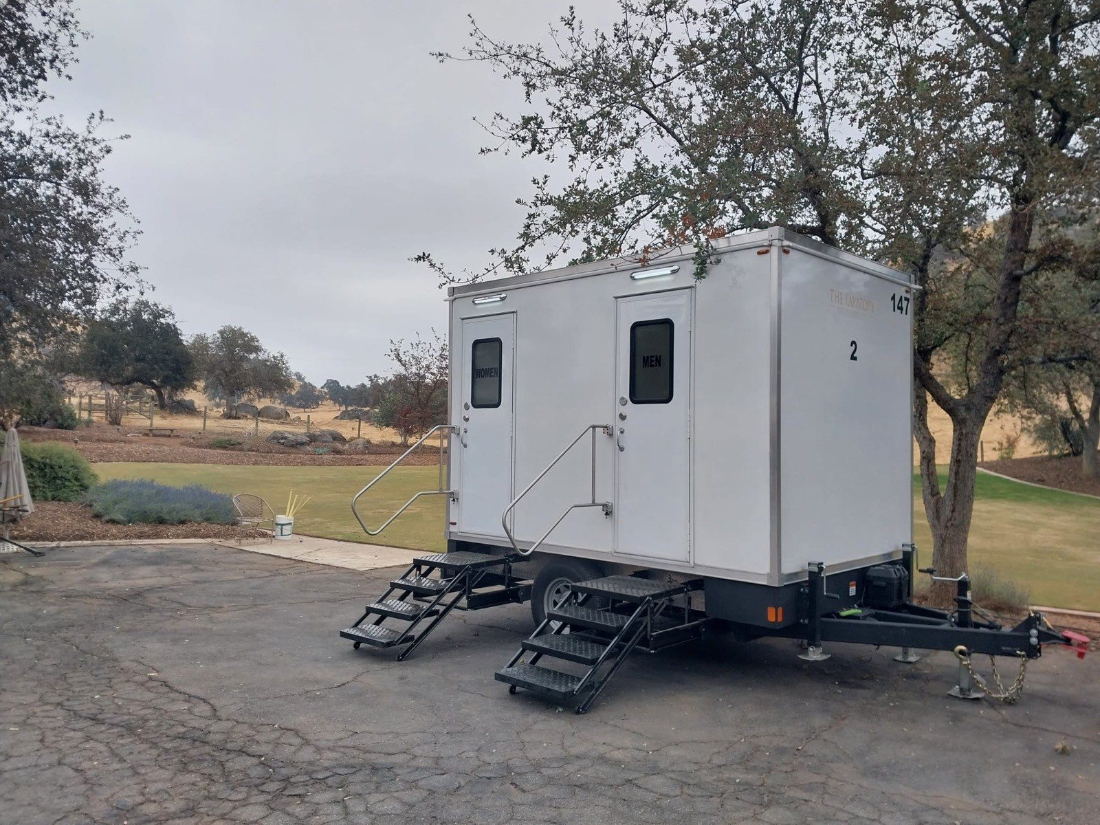A Lavatory 2-Station restroom trailer on a paved drive beside a golf course lawn, with oak trees and dry hills behind it