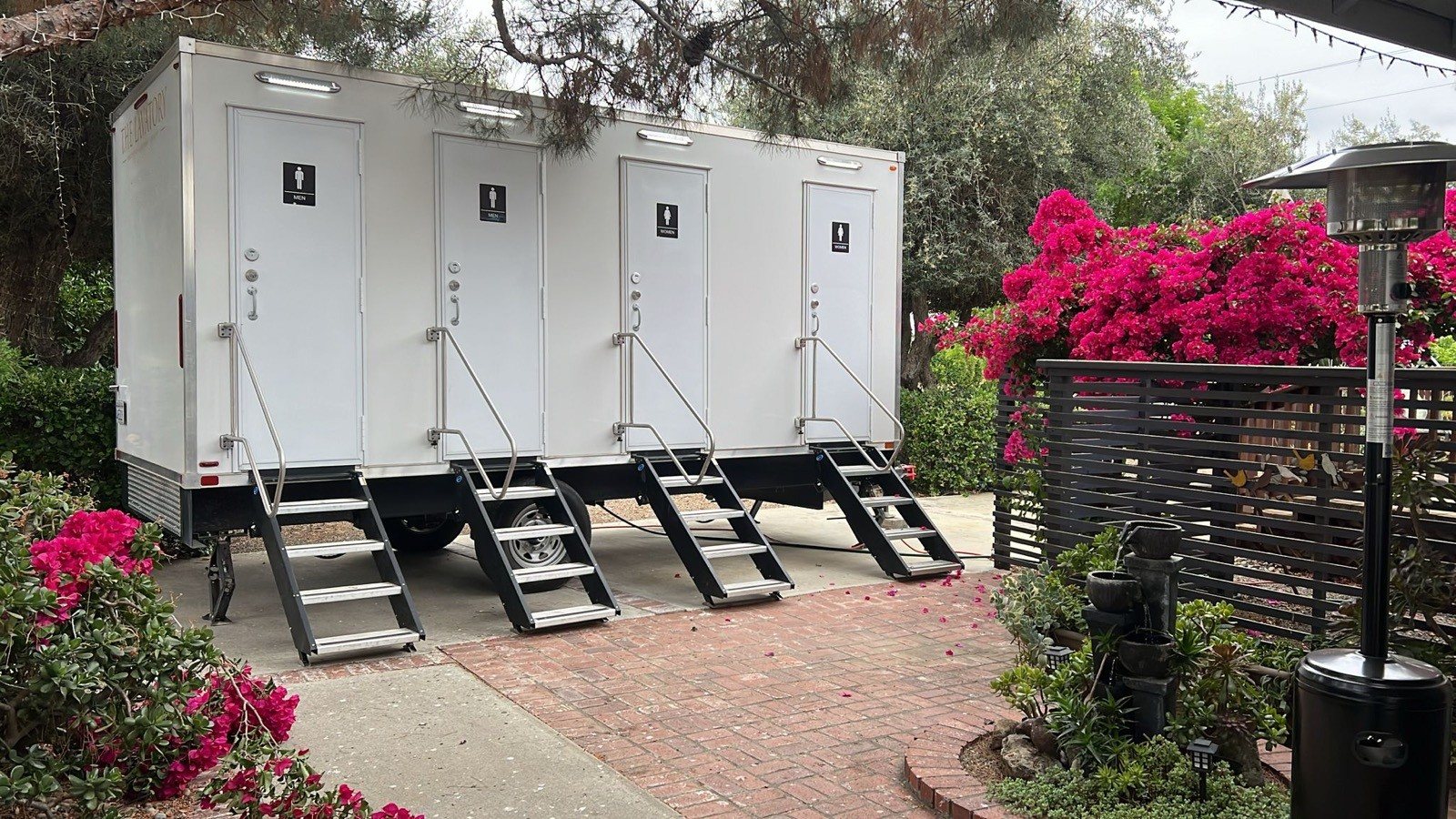 A Lavatory 4-Station restroom trailer on a brick garden patio beside pink bougainvillea and a patio heater