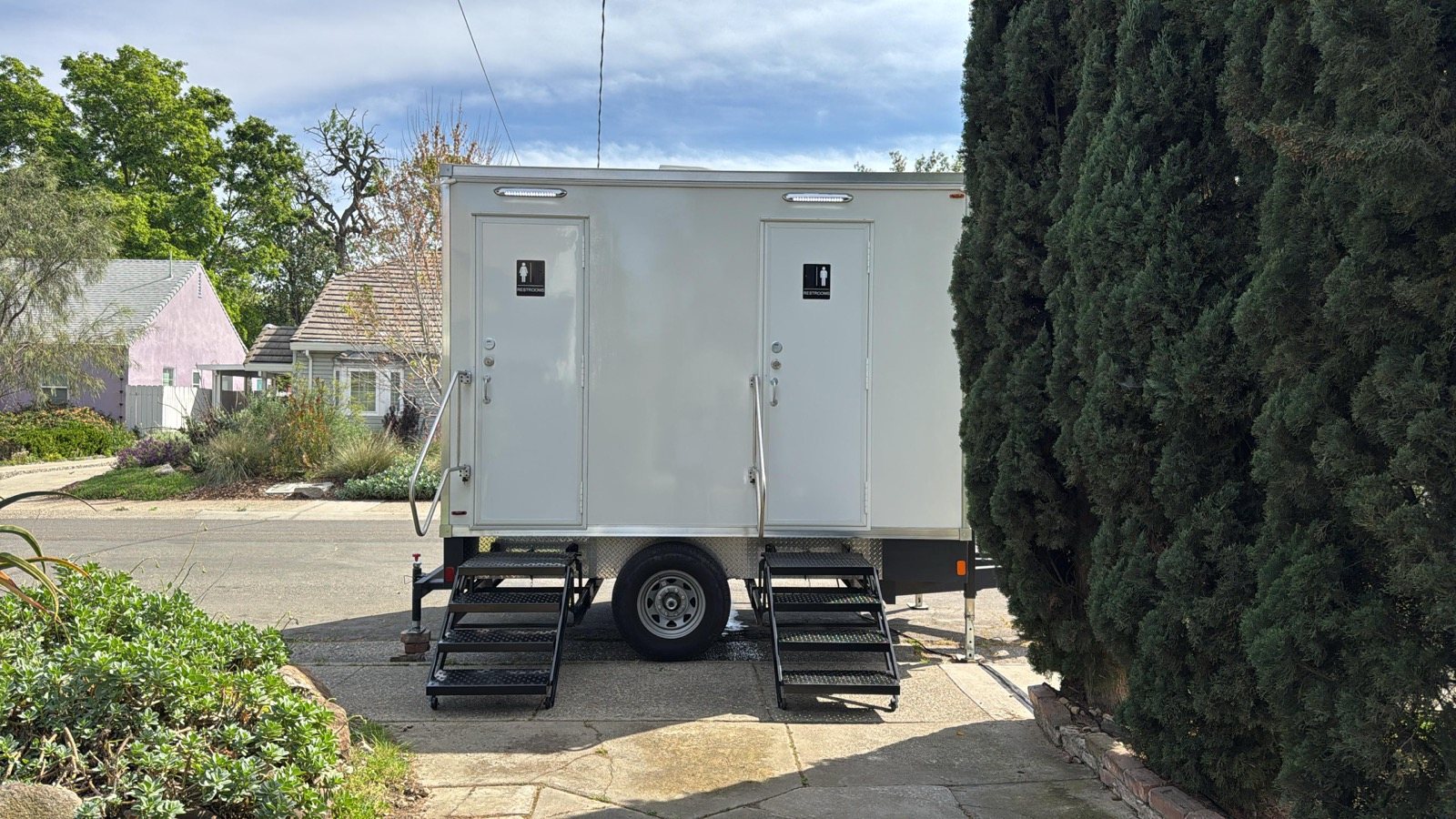 A Lavatory 2-Station restroom trailer parked on a residential drive beside a tall cypress hedge, with houses behind it