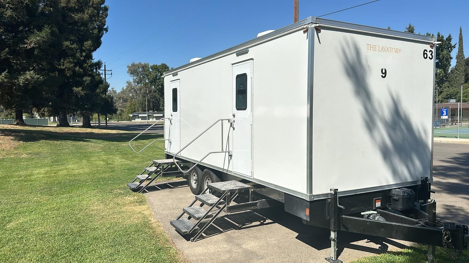 A Lavatory 9-Station restroom trailer on a paved pad at the edge of a green lawn, with shade trees and tennis courts behind it
