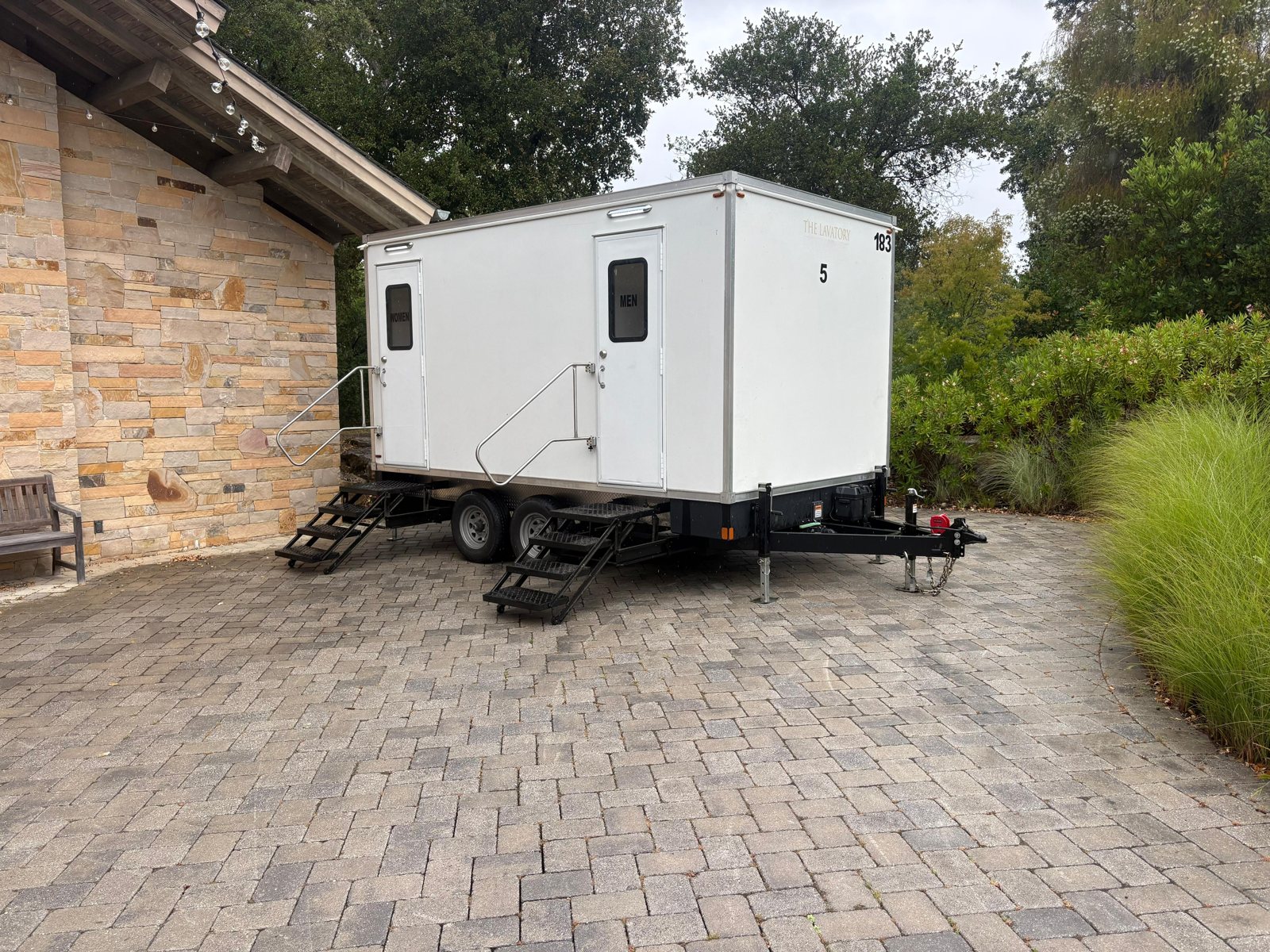 A Lavatory 5-Station luxury restroom trailer parked on a paver patio beside a stone building and landscaped garden