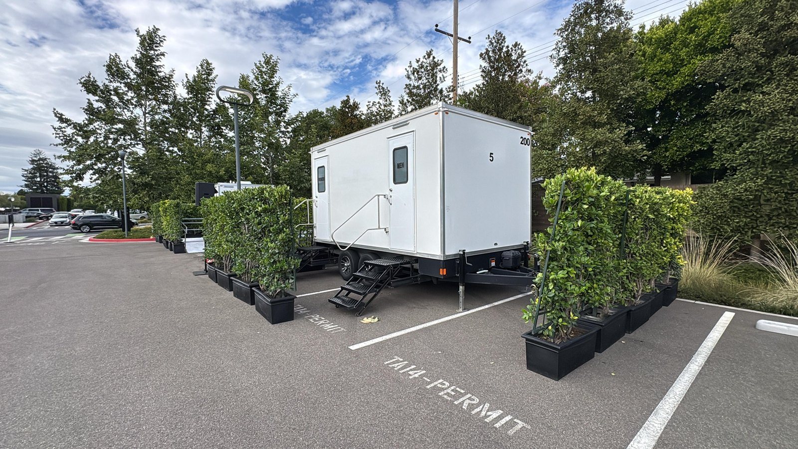 A Lavatory restroom trailer screened by rows of potted hedges in a parking lot set up for an event