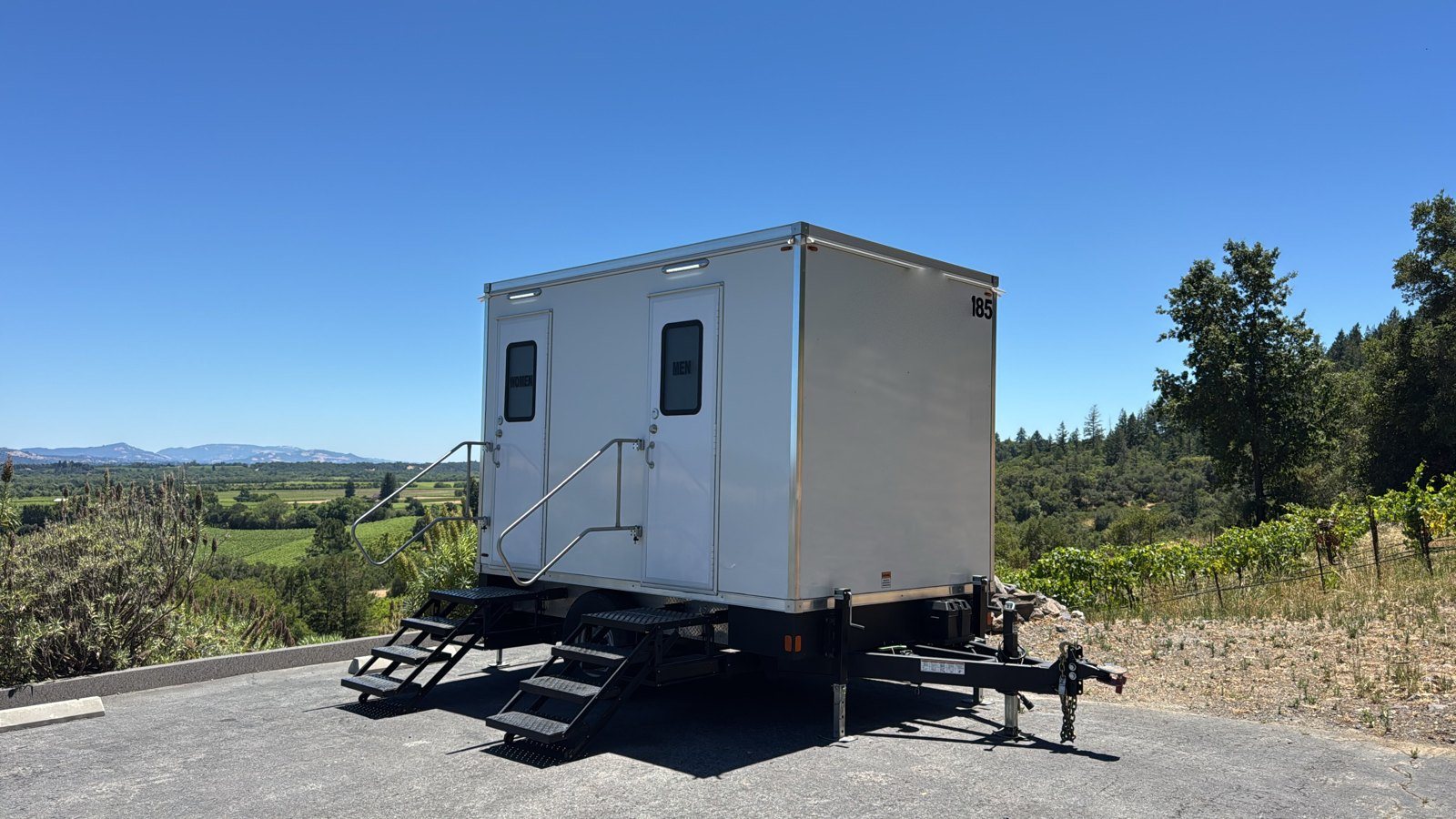 A Lavatory 2-Station restroom trailer on a paved pad at a hillside wedding site, with vineyard rows and mountains behind it