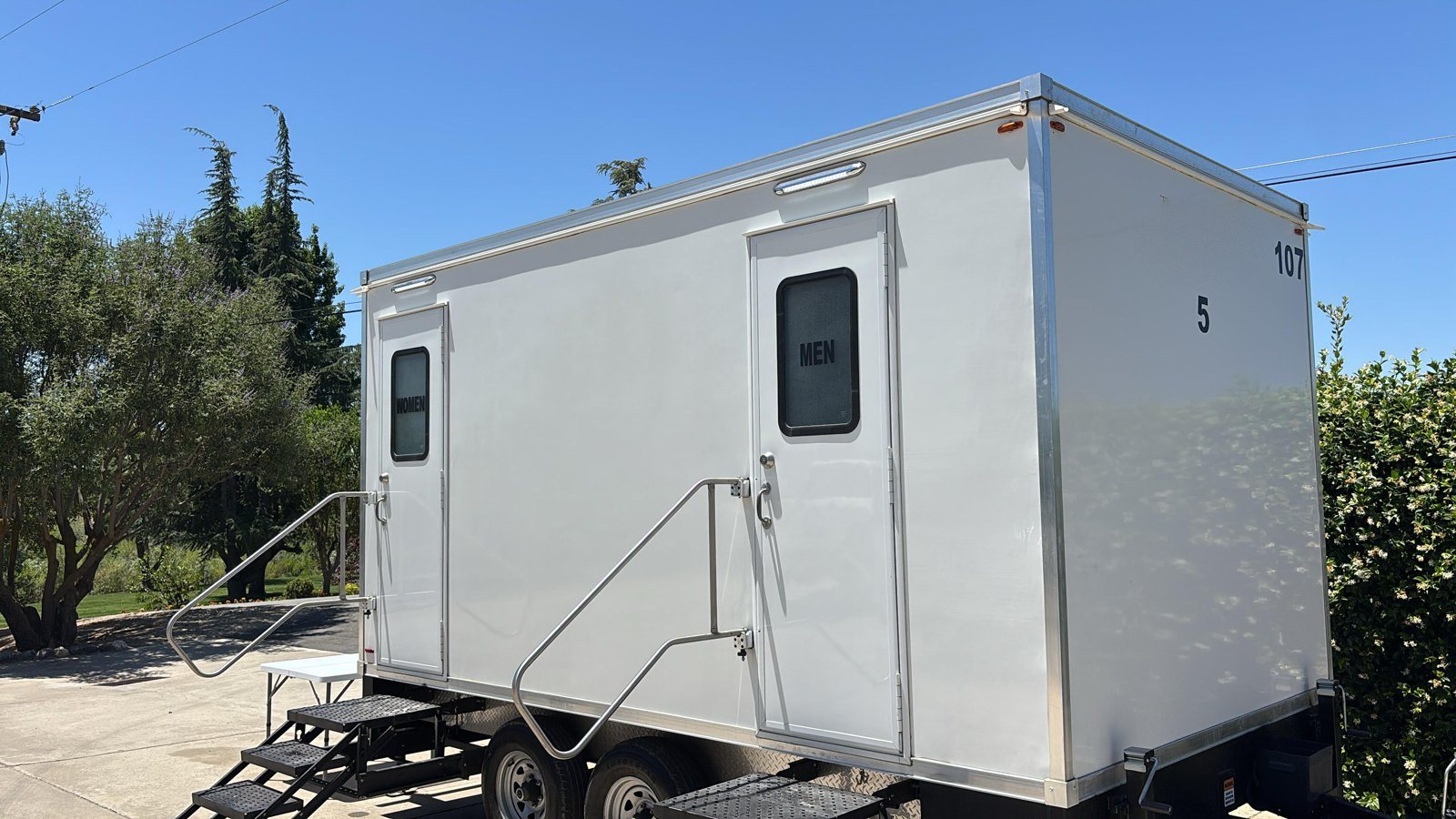 A Lavatory restroom trailer with steps and handrails parked on a paved residential drive beside olive trees and a hedge
