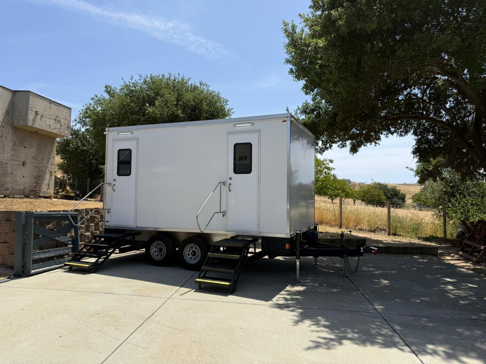 A Lavatory 5-Station luxury restroom trailer parked on a concrete pad under oak trees, with dry golden hills and a stone wall behind it