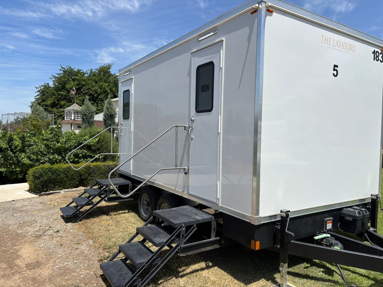 A Lavatory 5-Station luxury restroom trailer with two sets of stairs on a grass lawn beside a trimmed hedge under a blue sky