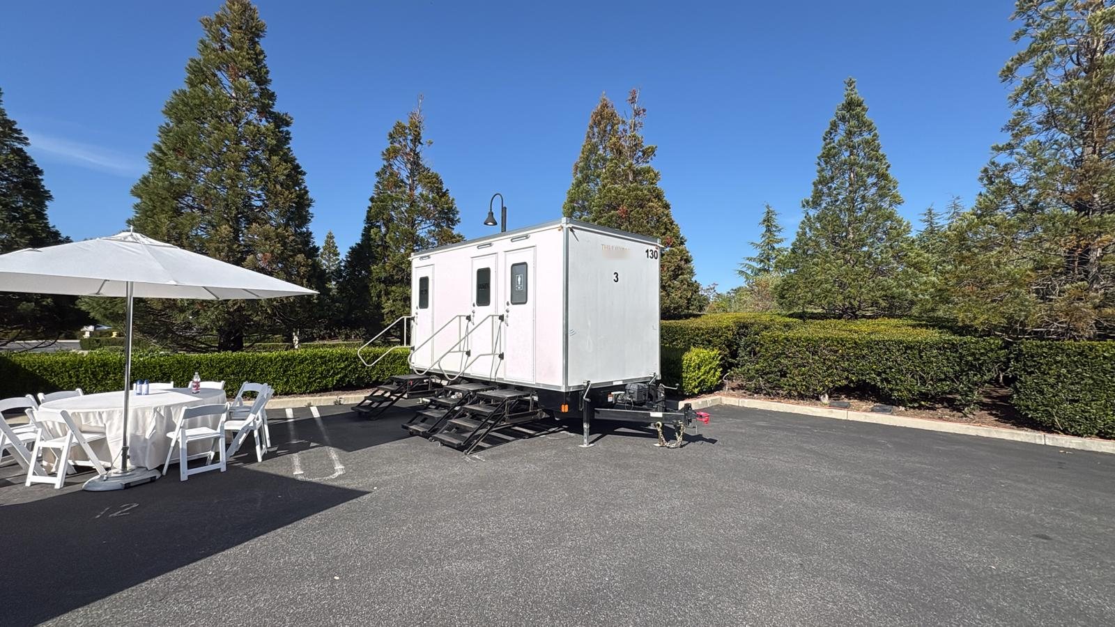 A three station Lavatory luxury restroom trailer parked on a paved event lot beside a white patio umbrella and folding chairs, with tall pines and a trimmed hedge behind it