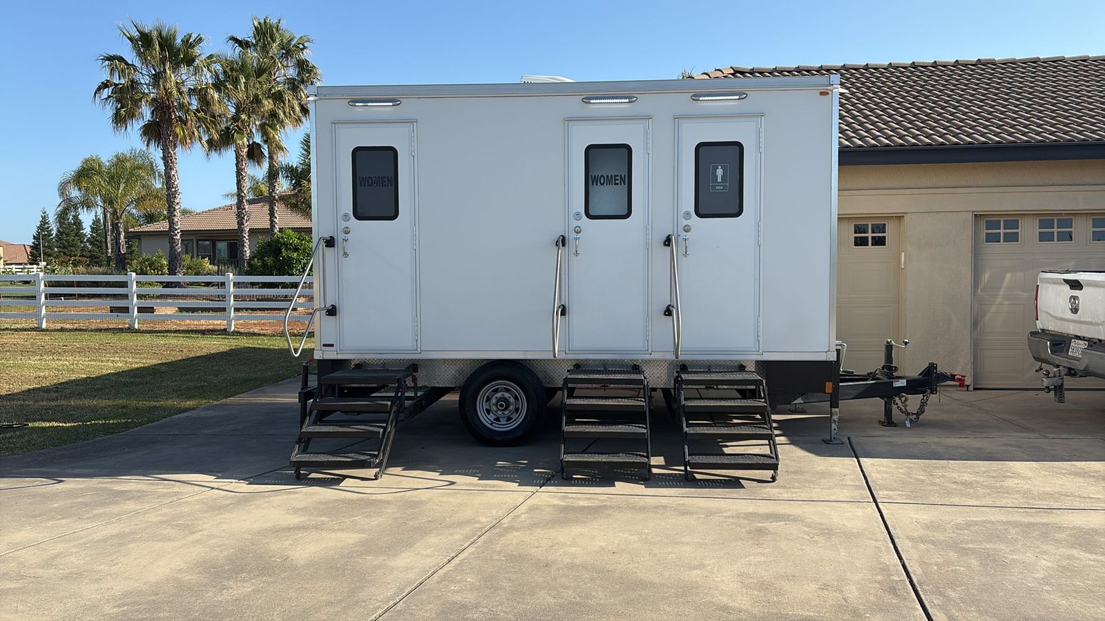 A Lavatory 3-Station restroom trailer on a home driveway with palm trees and a white rail fence behind it
