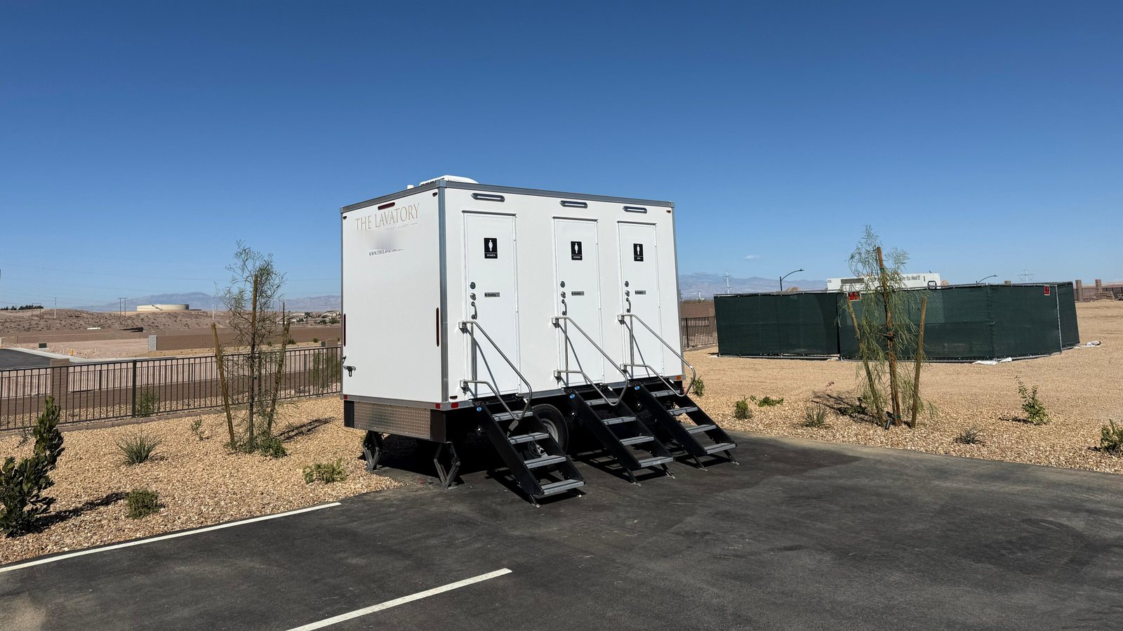 A Lavatory 3-Station restroom trailer on a paved lot with desert landscaping, dry ground and distant mountains under a clear sky