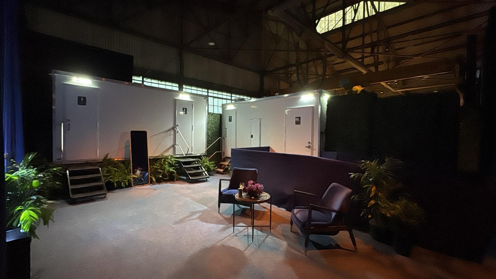 Two Lavatory restroom trailers lit up inside a large warehouse event space with lounge chairs in front