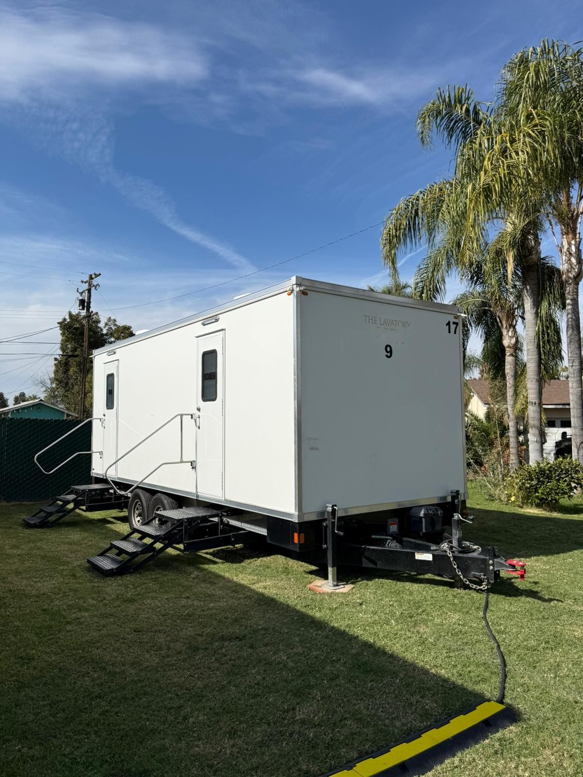 A Lavatory restroom trailer on a green lawn with palm trees behind it