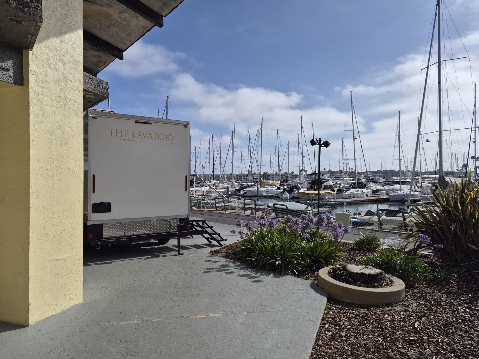 A Lavatory restroom trailer beside a building at a marina, with sailboat masts in the background