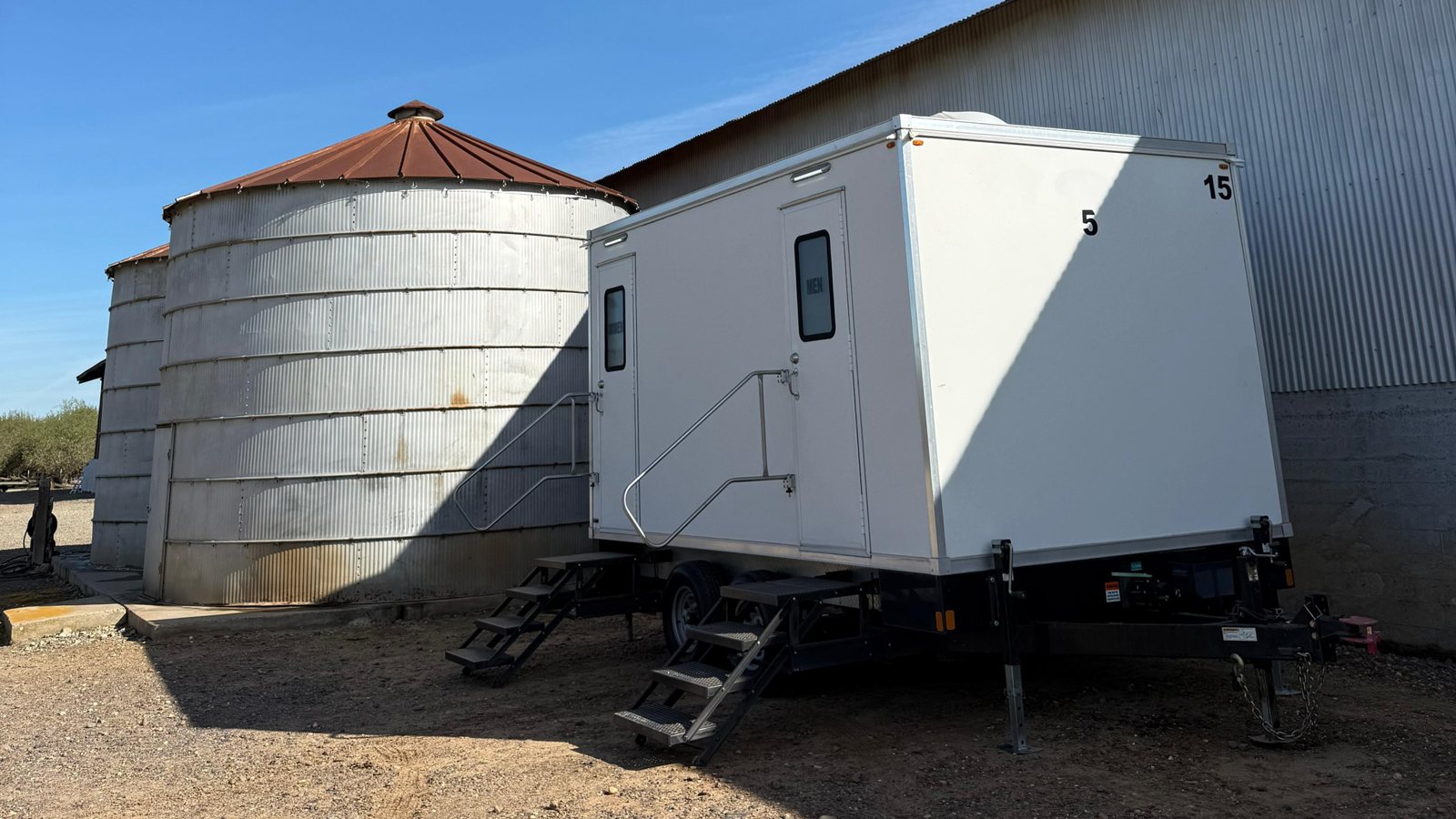 A Lavatory 5-Station luxury restroom trailer with steel stairs and handrails parked beside a grain silo and a metal farm building in Merced