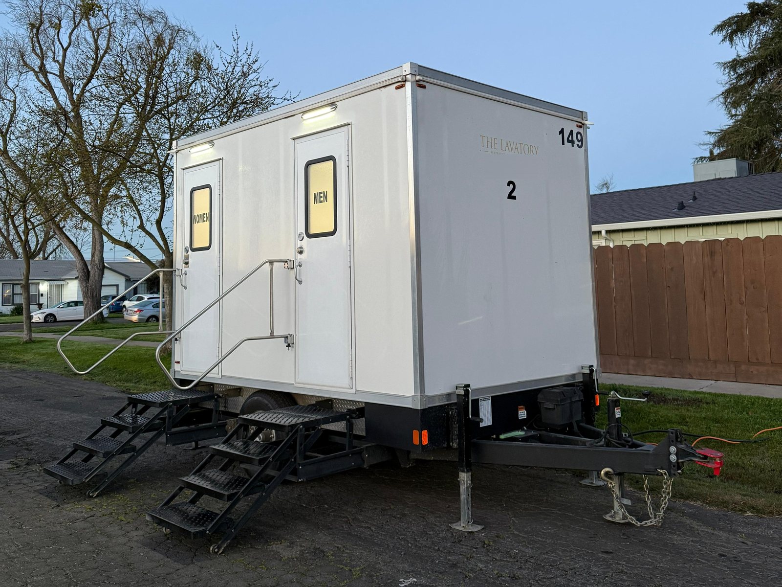 A Lavatory 2-Station restroom trailer with its door lights on, parked on a residential street in Merced at dusk