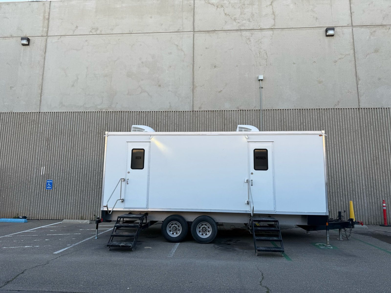 A Lavatory 9-Station restroom trailer with men's and women's doors parked in a paved lot beside a large concrete building in Merced