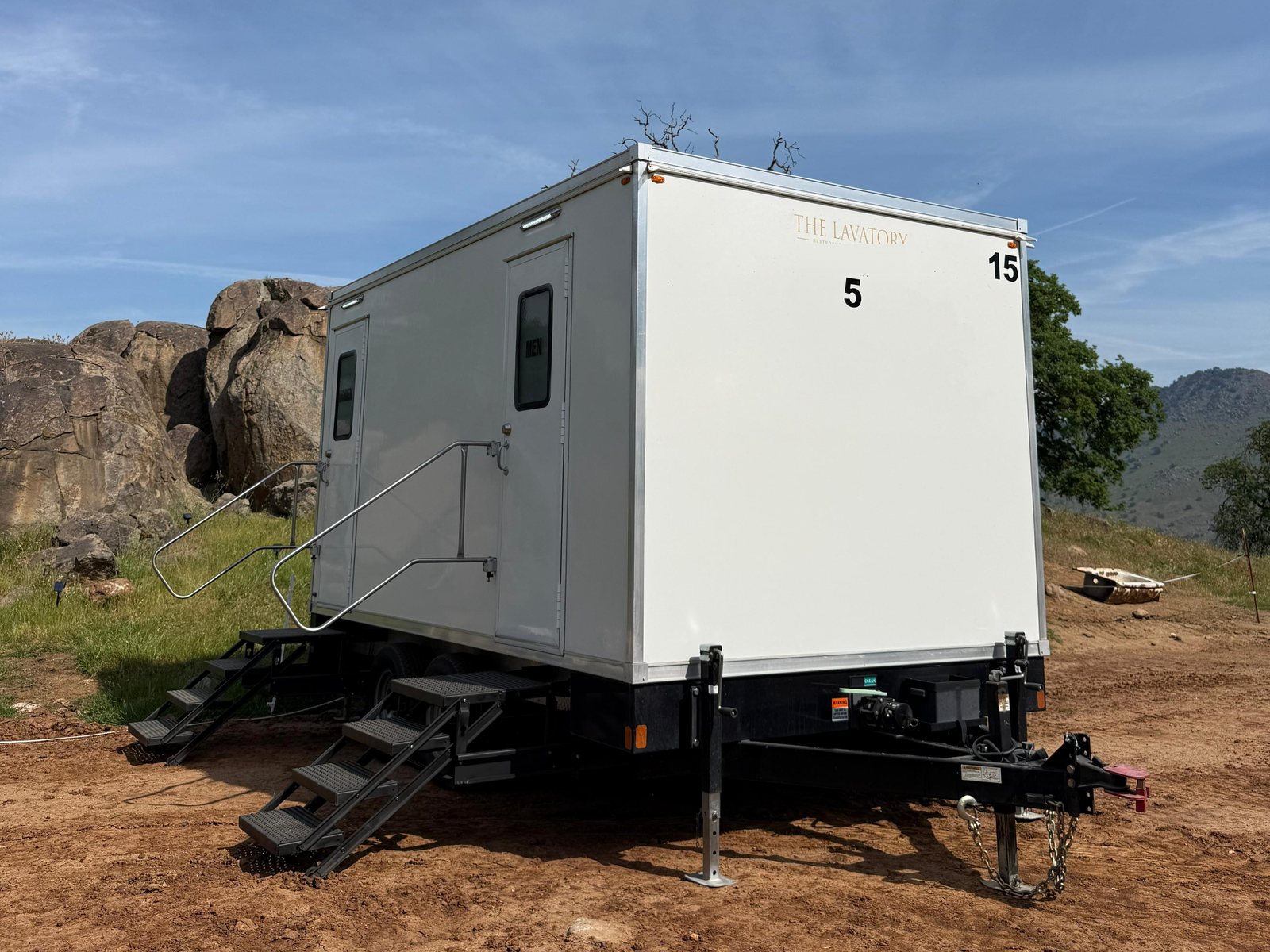 A Lavatory 5-Station restroom trailer on leveling jacks on a dirt pad beside granite boulders and oak-covered foothills
