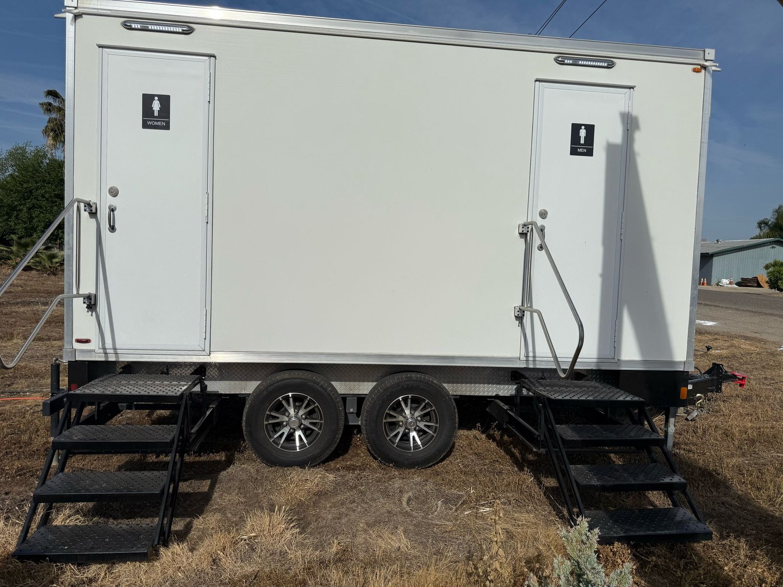 A Lavatory 5-Station restroom trailer with separate women's and men's doors and steel stairs set up on dry grass at a rural property