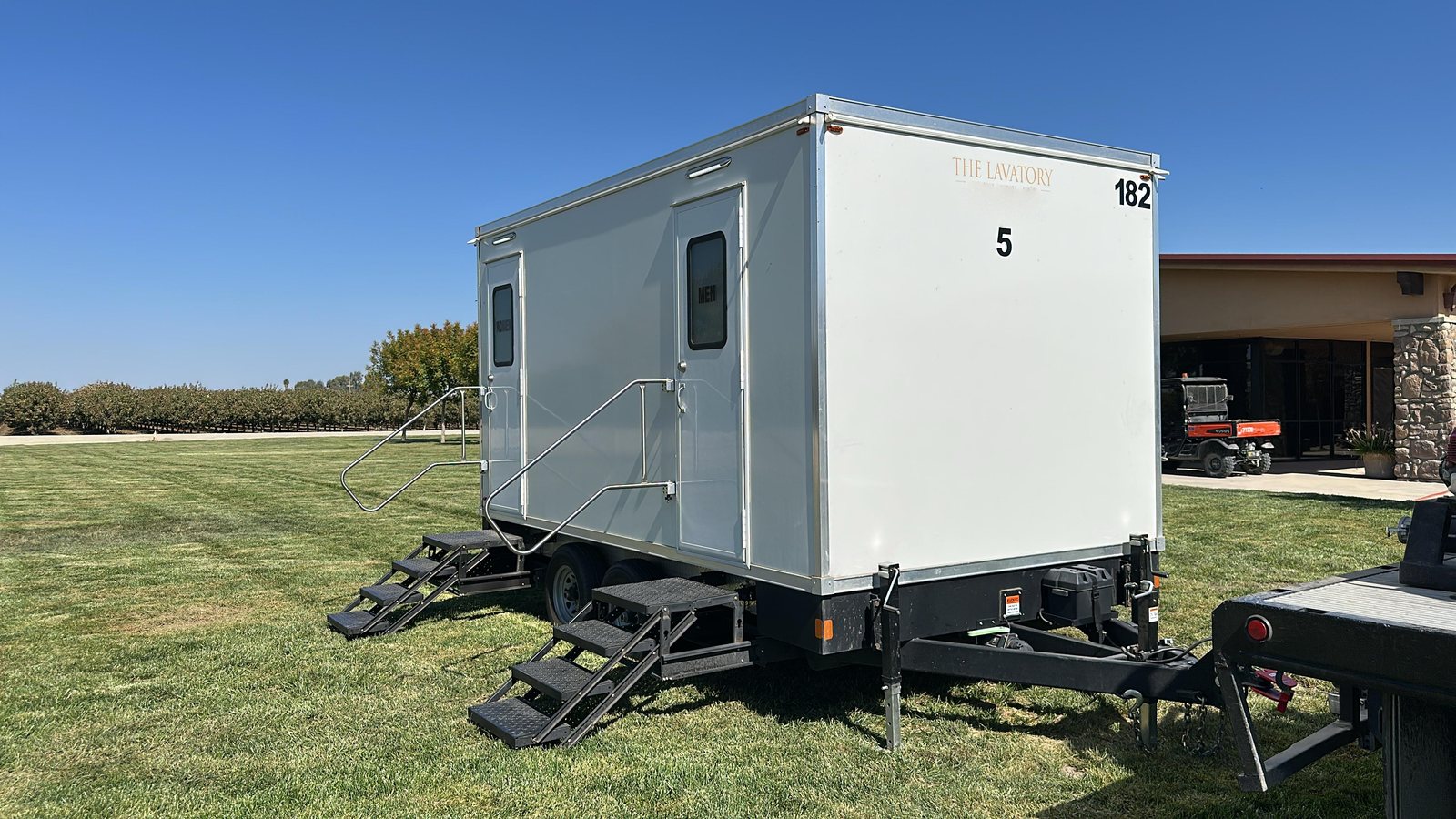 A Lavatory 5-Station restroom trailer on a mowed lawn with an orchard row and a low building behind it