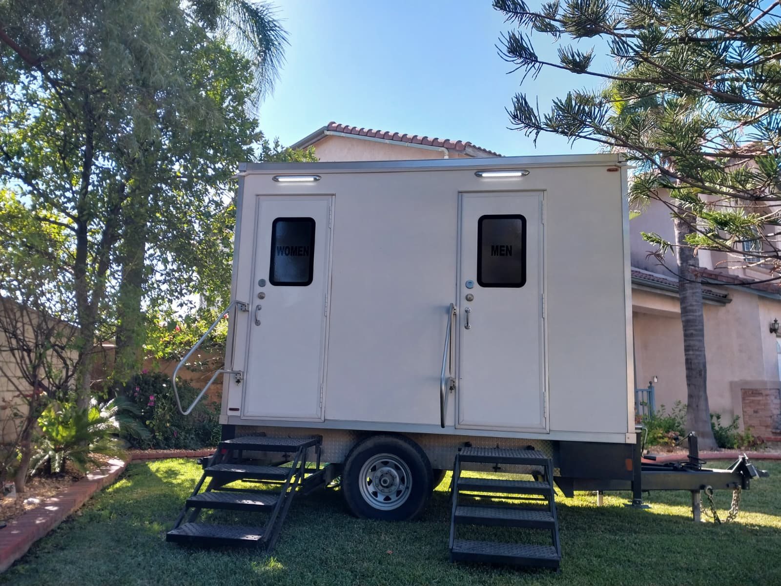 A two station Lavatory luxury restroom trailer set on a backyard lawn beside a two story stucco home, with palm and pine trees overhead