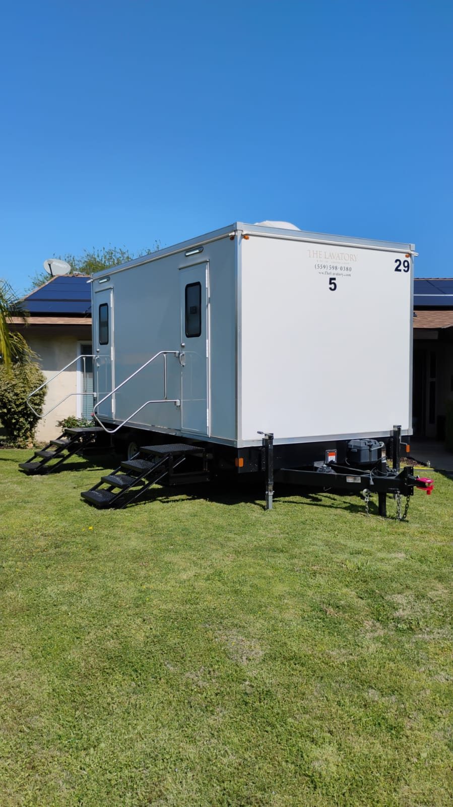 A Lavatory 5-Station restroom trailer parked on a green lawn beside a single story house on a sunny day