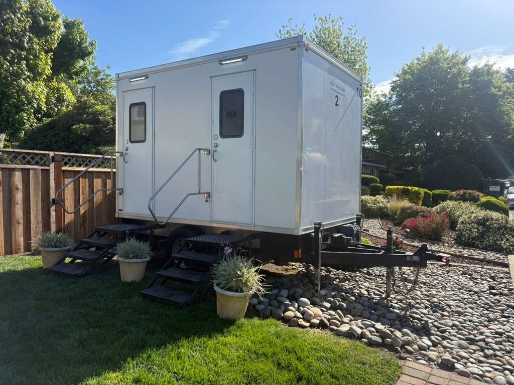 A two station Lavatory restroom trailer set on a backyard lawn beside a wood fence, with potted plants at the steps