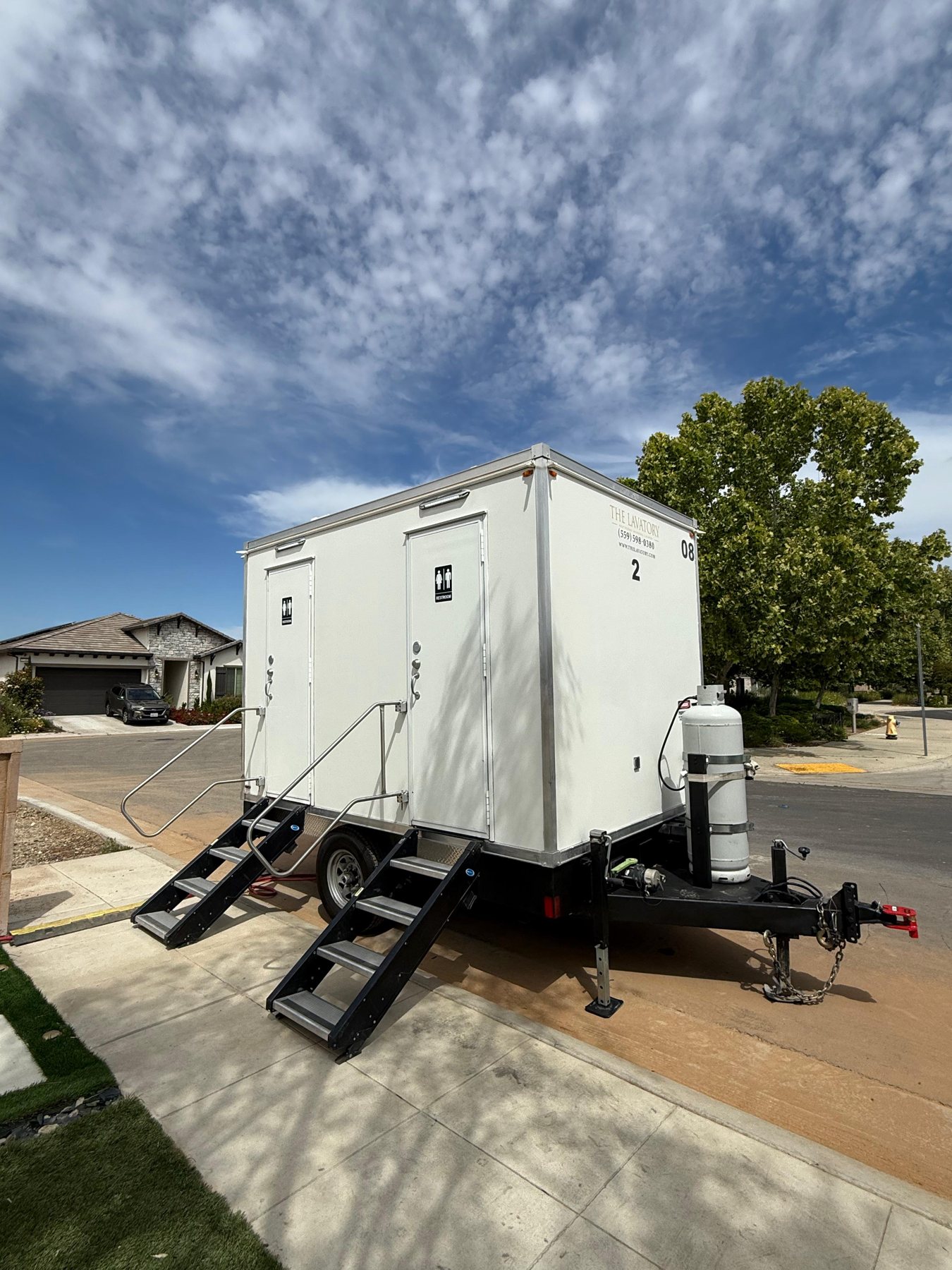A two station Lavatory restroom trailer parked on a home driveway with a tile roof house behind it and a generator beside the tongue