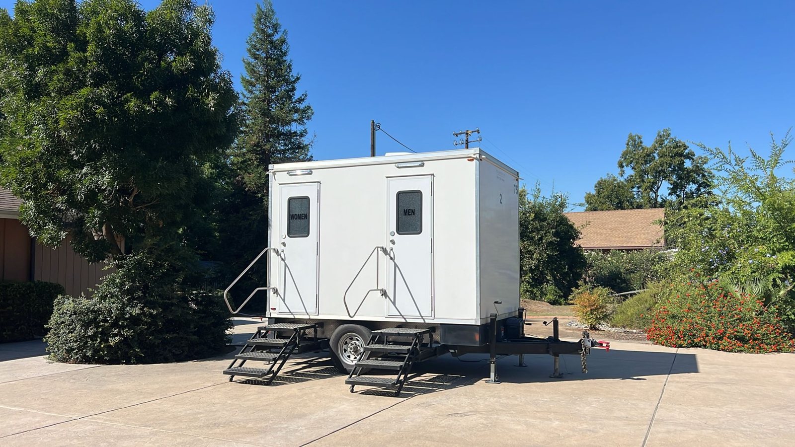 A two station Lavatory restroom trailer parked on a wide paved drive in front of a home, surrounded by shrubs and trees