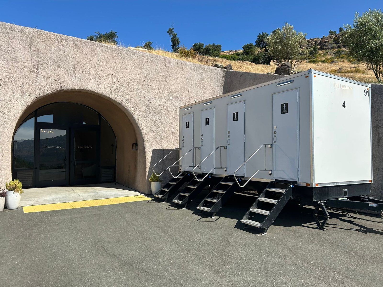 A Lavatory four-door luxury restroom trailer with steel stairs and handrails parked beside a stucco winery cave entrance below a dry hillside with oaks