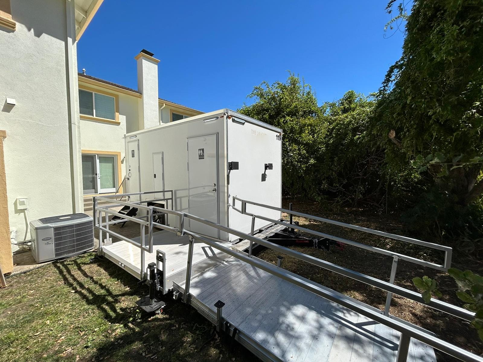 A Lavatory ADA restroom trailer with a long aluminum ramp and handrails set on the side lawn of a two-story home