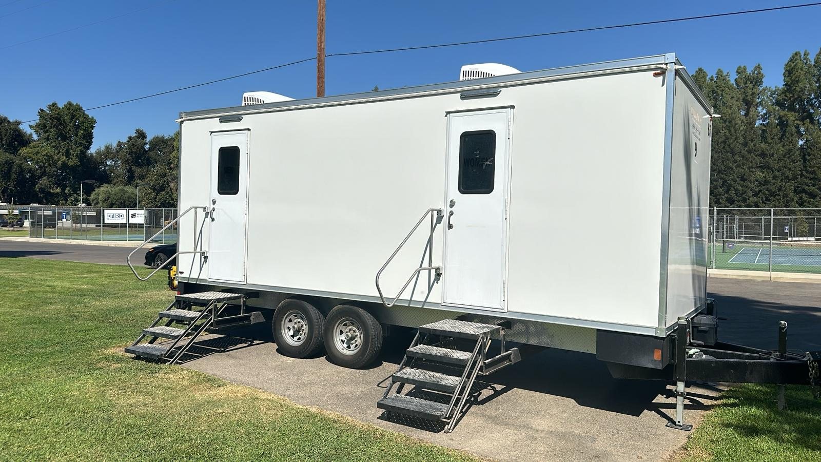 A Lavatory 9-Station restroom trailer with two entry stairs parked on a park lawn next to fenced tennis courts