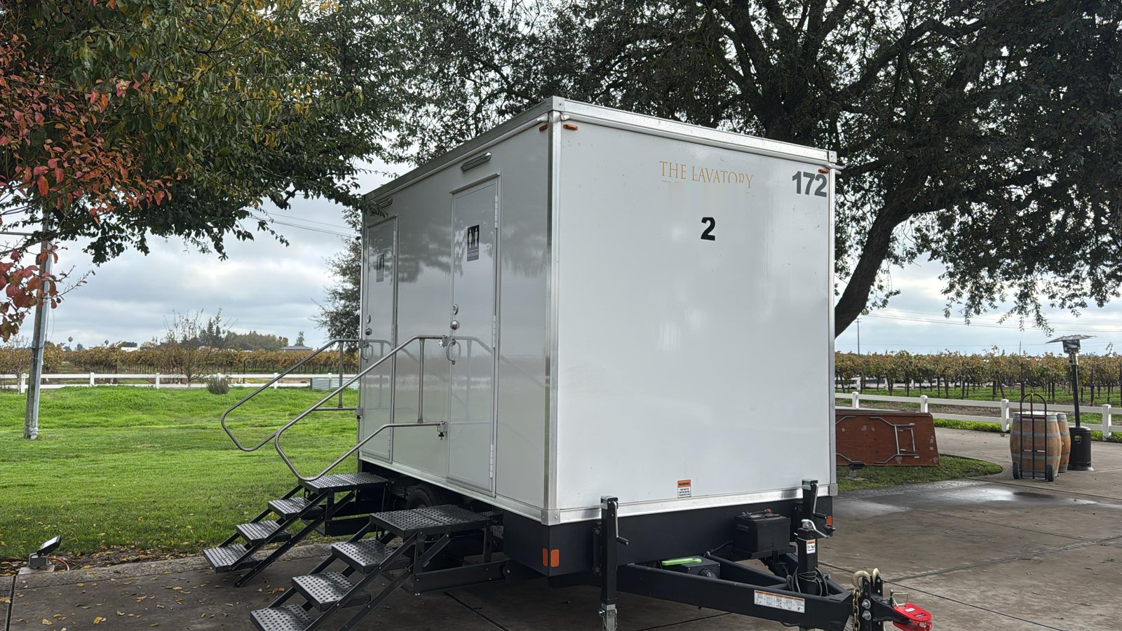 A Lavatory 2-Station restroom trailer on a concrete pad under a large oak, with vineyard rows, a white rail fence and wine barrels behind it