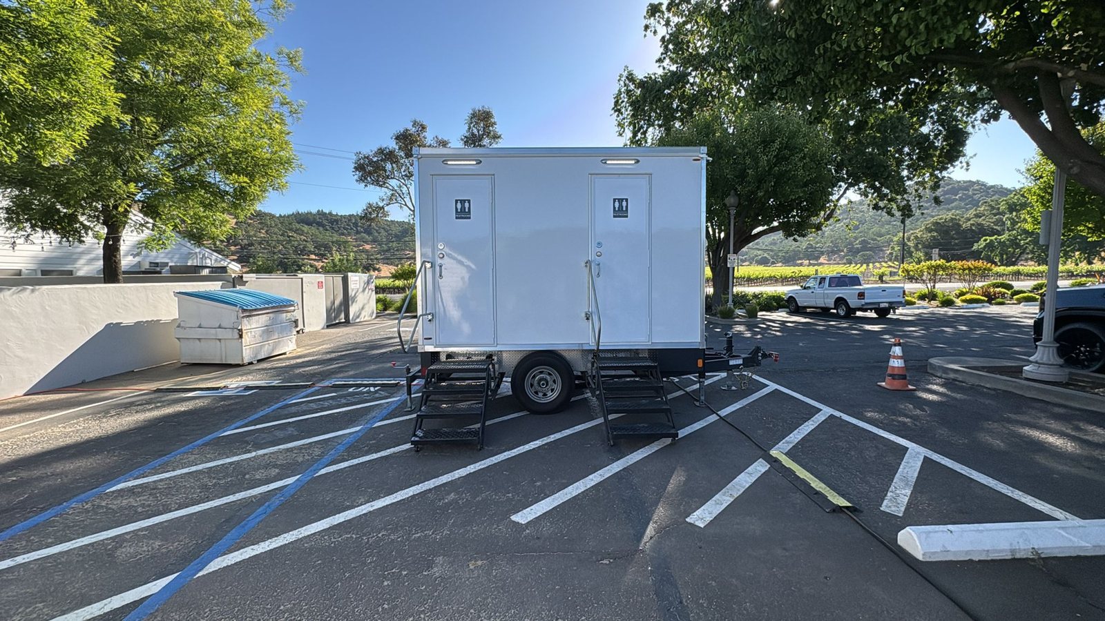 A Lavatory 2-Station restroom trailer parked in a winery lot with vineyard rows and wooded hills behind it