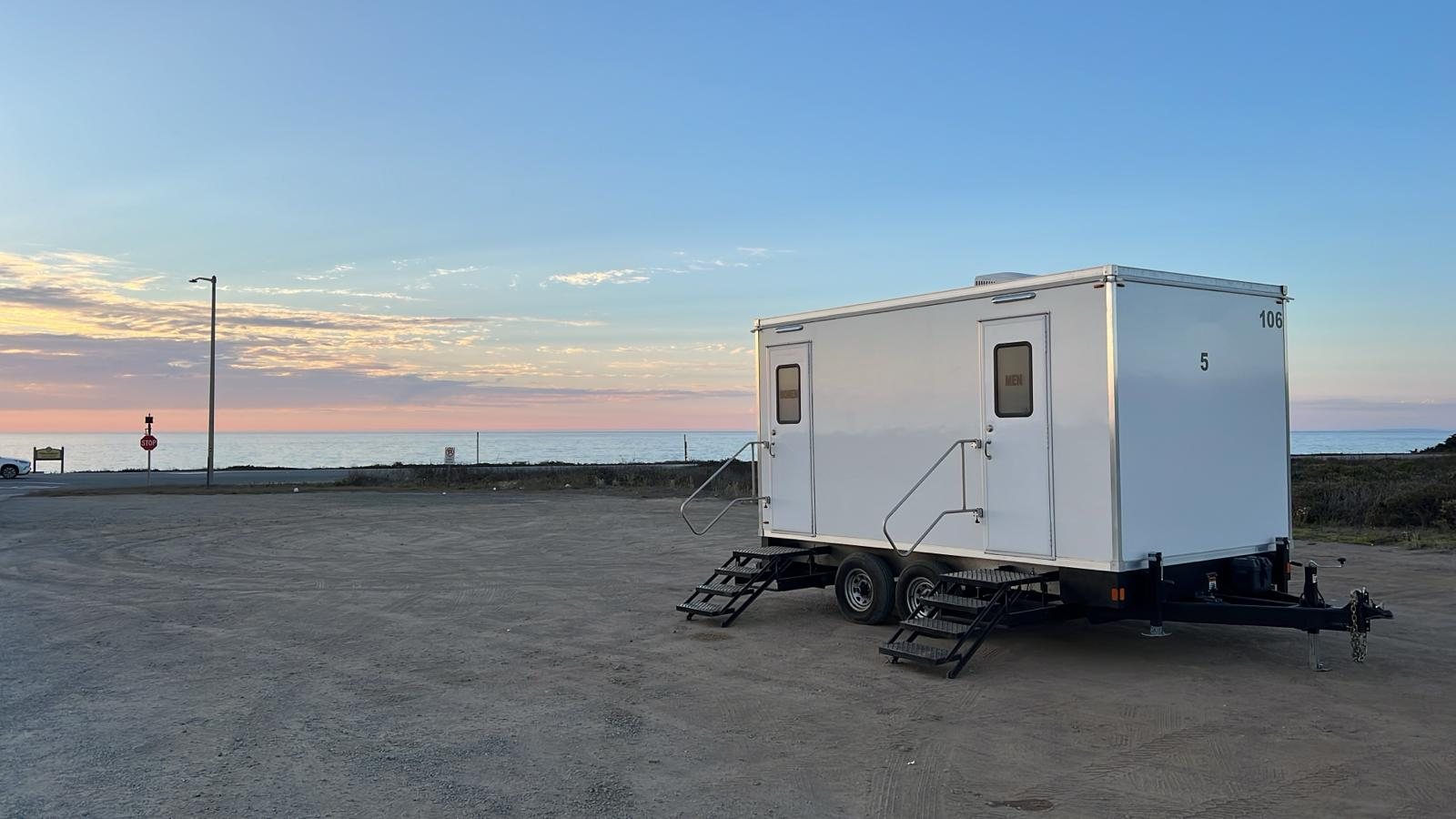 A Lavatory 5-Station luxury restroom trailer parked on a gravel lot by the coast at sunset, with the ocean on the horizon