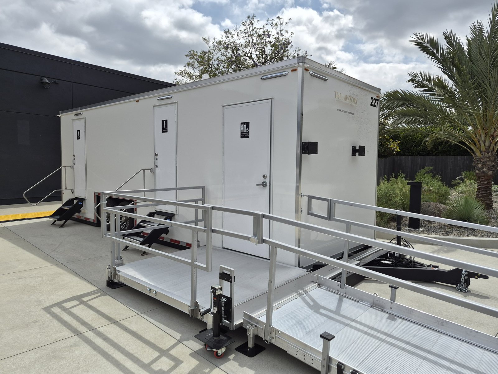 A Lavatory ADA + 2 restroom trailer with a long ramp and railings set up in a commercial courtyard with palm trees