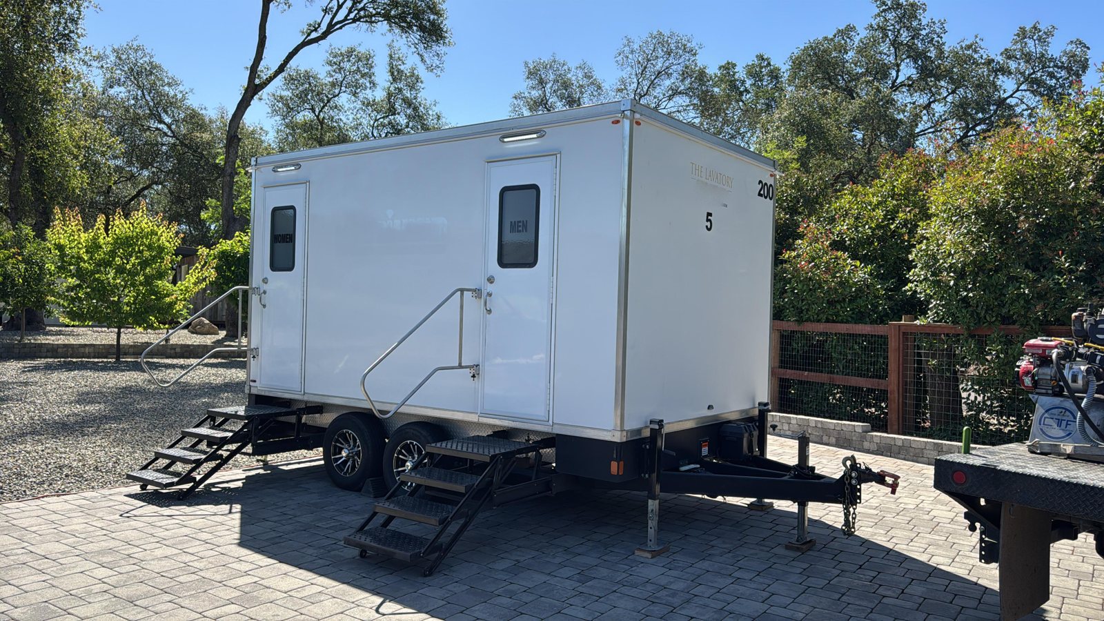 A Lavatory 5-Station restroom trailer on a paver drive beside a wooden fence and shade trees