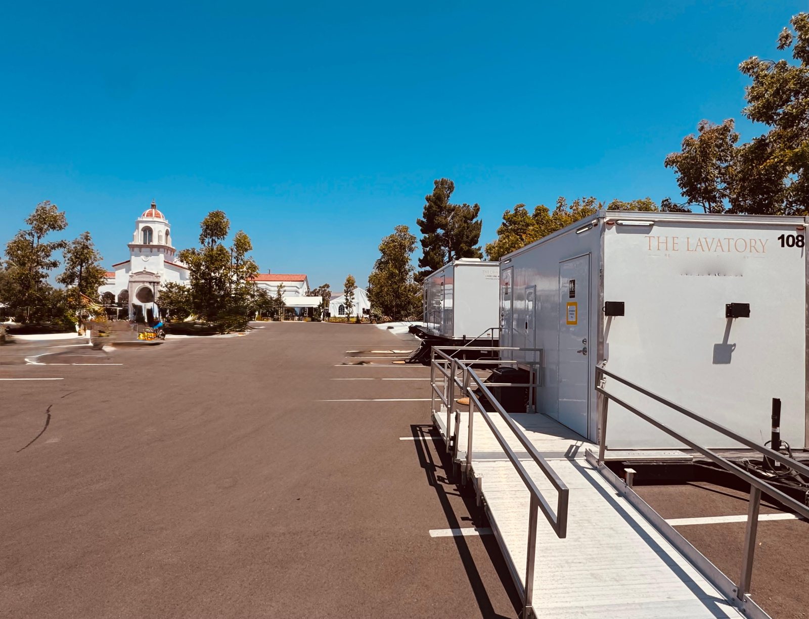 Two Lavatory restroom trailers with ramps and railings parked in a church parking lot beside a building with a bell tower