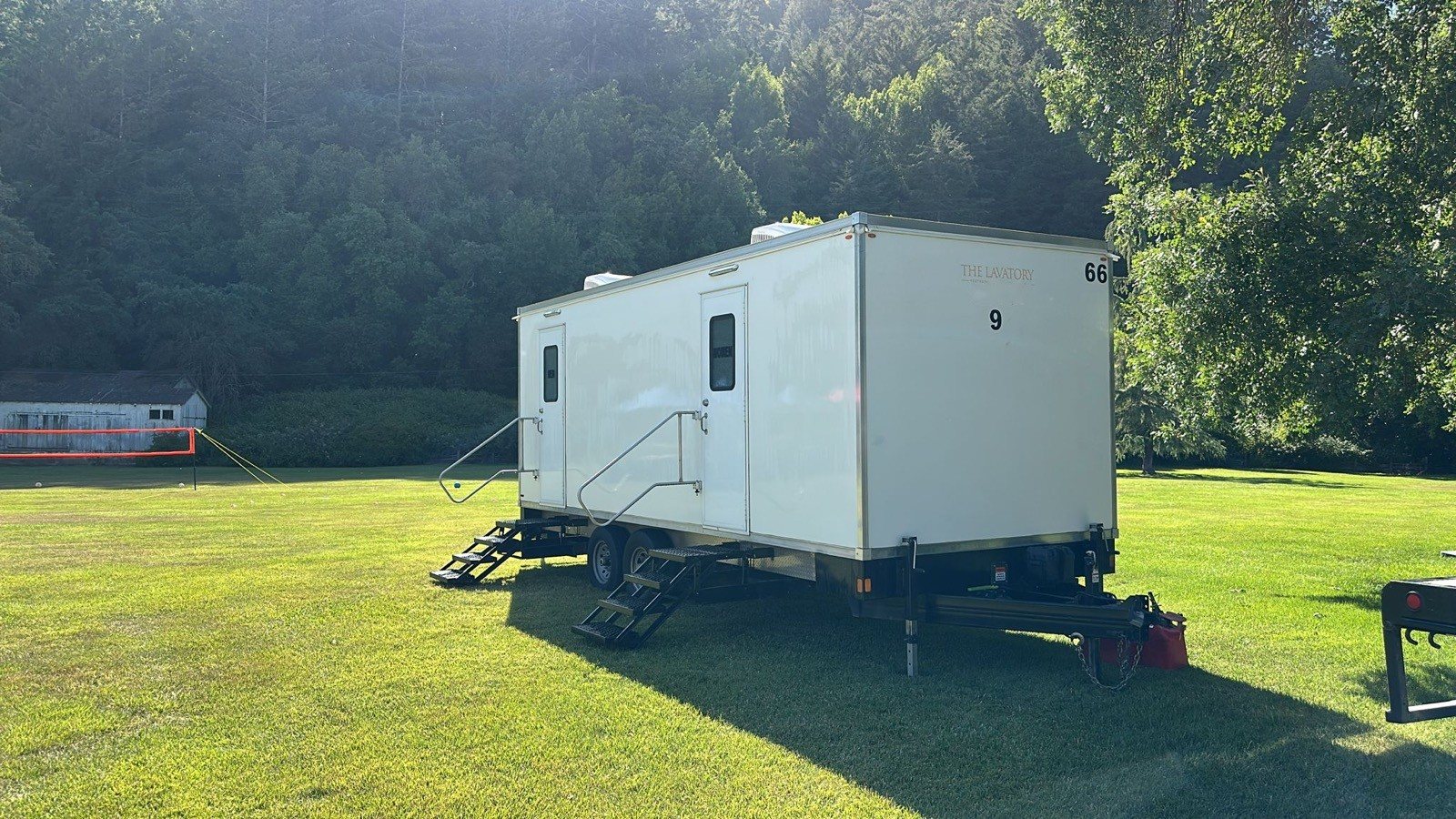 A Lavatory 9-Station restroom trailer on a sunny lawn with a tree-covered hillside behind it